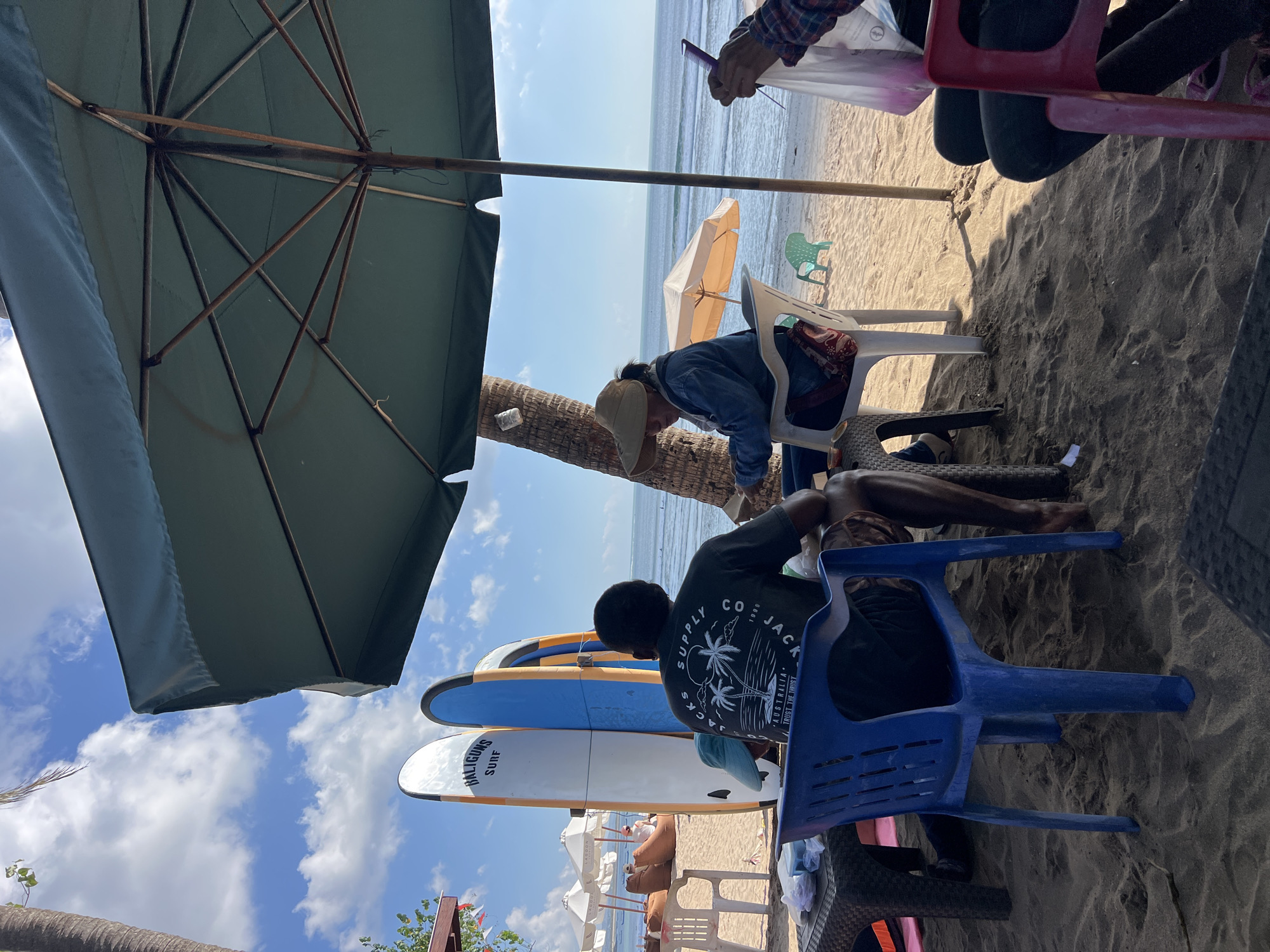 At a beachside cafe in Kuta, Indonesia, two individuals are seated under a large green umbrella, interacting with each other. The man in the foreground, wearing a dark t-shirt with a palm tree graphic, is seated in a blue plastic chair, facing away from the camera. A woman, wearing a wide-brimmed hat and a denim jacket, sits opposite him in a white plastic chair, looking down at her hands. She appears to be holding a phone or tablet.
In the background, three surfboards stand upright on the sand, labeled "BALIGUNA SURF". Further back, rows of white beach umbrellas and beanbag chairs are set up on the sand, indicating a tourist area. The ocean stretches to the horizon under a bright, partly cloudy sky. The sand is a light tan color, and the scene is bathed in natural sunlight, casting shadows from the umbrella and palm trees. The overall atmosphere suggests a casual, relaxed setting in a popular Indonesian beach resort town.