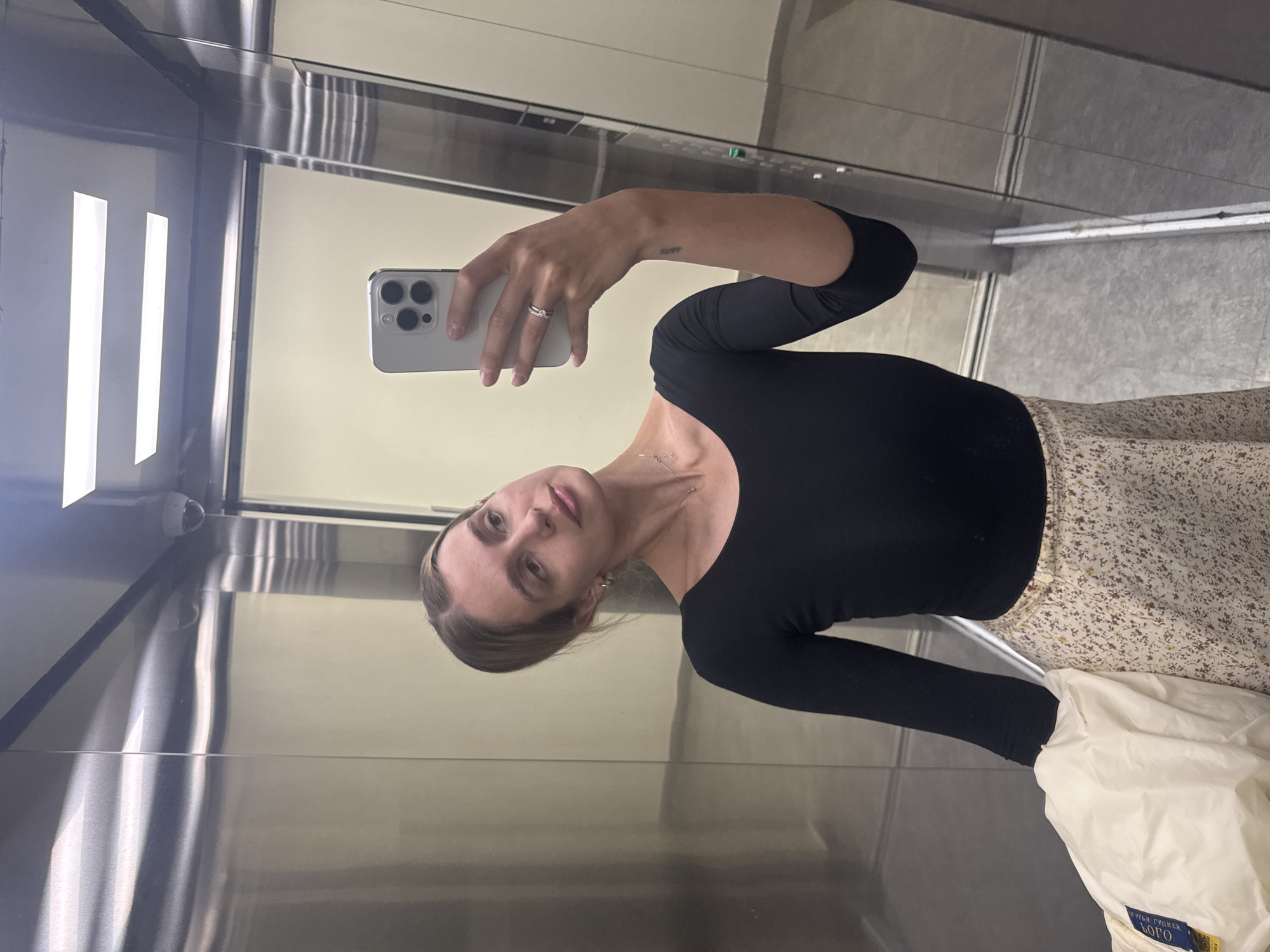 A young woman is captured taking a mirror selfie inside an elevator, the image rotated 90 degrees clockwise. She is positioned slightly to the right of the frame, holding a light-colored smartphone with her right hand, which has a silver ring on her ring finger. Her gaze is directed towards the camera. She has light brown hair pulled back, exposing her face, and is wearing a form-fitting black long-sleeve top. A delicate silver necklace is visible around her neck, and a small, stylized tattoo reading "XURY" is visible on her right forearm, just above the wrist. Her lower body is clad in a light-colored skirt with a subtle floral or speckled pattern. She carries a white, possibly puffy, bag or jacket draped over her left arm, with some blue branding that includes Cyrillic text, appearing to read "КУБИ КУБЕЙ ЛОГО".
The setting is an elevator with highly reflective, metallic walls, likely stainless steel, which act as mirrors. The reflections create a sense of depth and show parts of the elevator interior, including linear light fixtures. The floor appears to be a light gray, possibly tiled or textured. In the top left corner of the elevator's reflection, a small security camera is visible. The lighting is artificial, likely from within the elevator, creating bright reflections. The overall scene suggests an indoor environment, with no discernible weather or time of day cues other than artificial illumination.