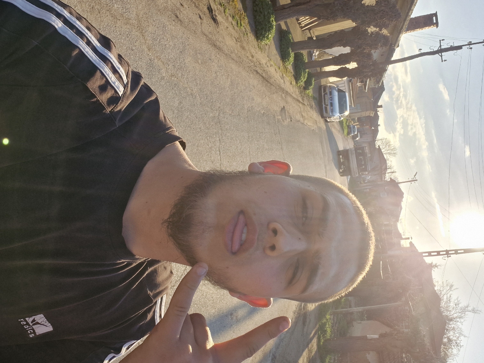 A man is posing for a selfie on a street in Hirevo, Bulgaria. The image captures him from the chest up, with his head tilted to the side, sticking his tongue out slightly. His right hand is raised with his index and pinky fingers extended, making the "rock on" gesture. He has short hair and a beard. He is wearing a dark t-shirt with white stripes on the shoulder and a small logo on the lower left.
The street appears to be a quiet residential road with trees and buildings lining it. In the background, there are parked cars and houses. Power lines are visible above the buildings. The sunlight is bright, suggesting it is late afternoon or early evening. The image is slightly overexposed.