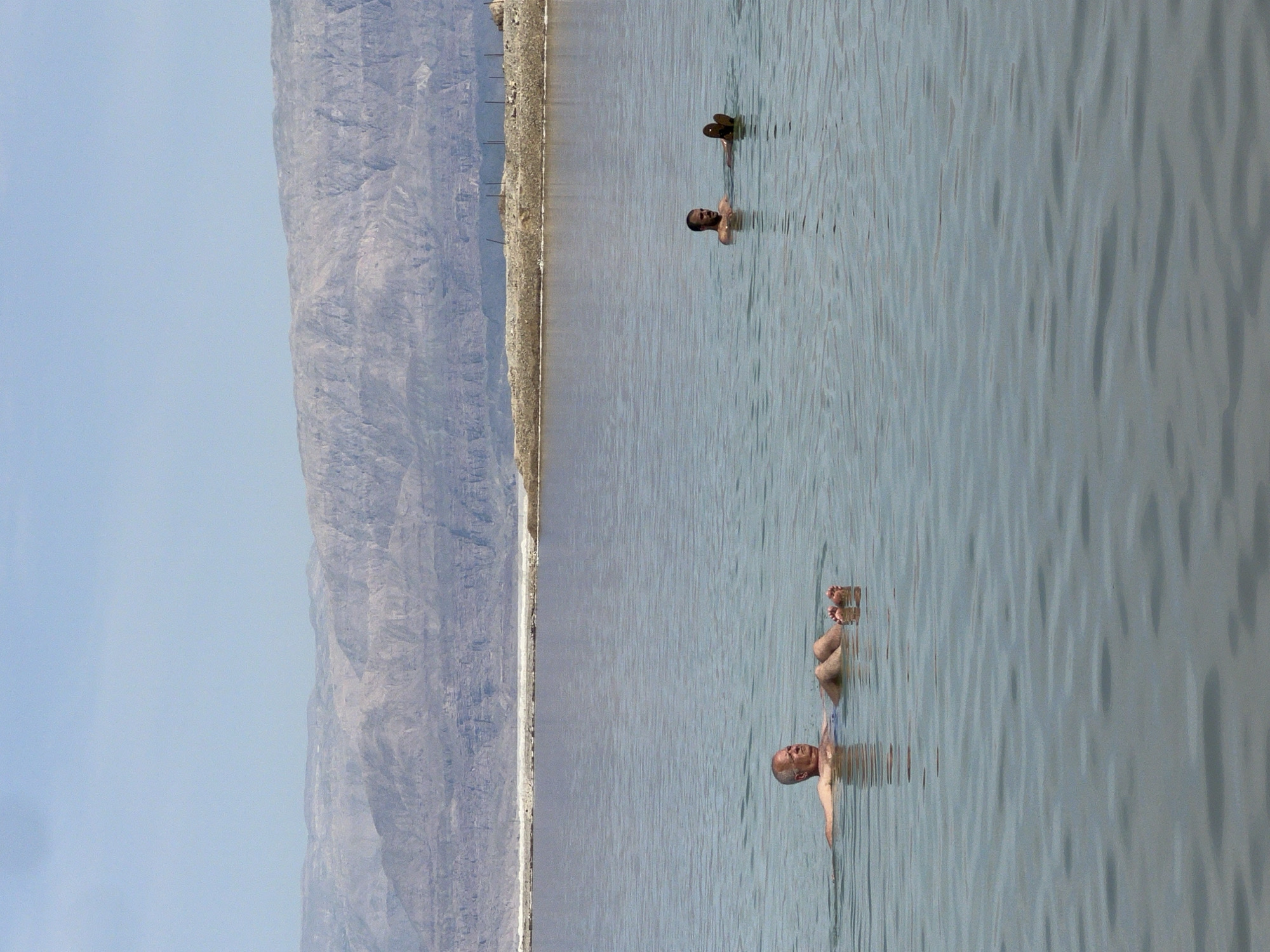 The photograph depicts the Dead Sea in the Tamar Regional Council, Israel. Two individuals are seen floating in the water, one with their feet above the surface. A mountain range stretches along the left side of the frame. The sky is a pale blue.