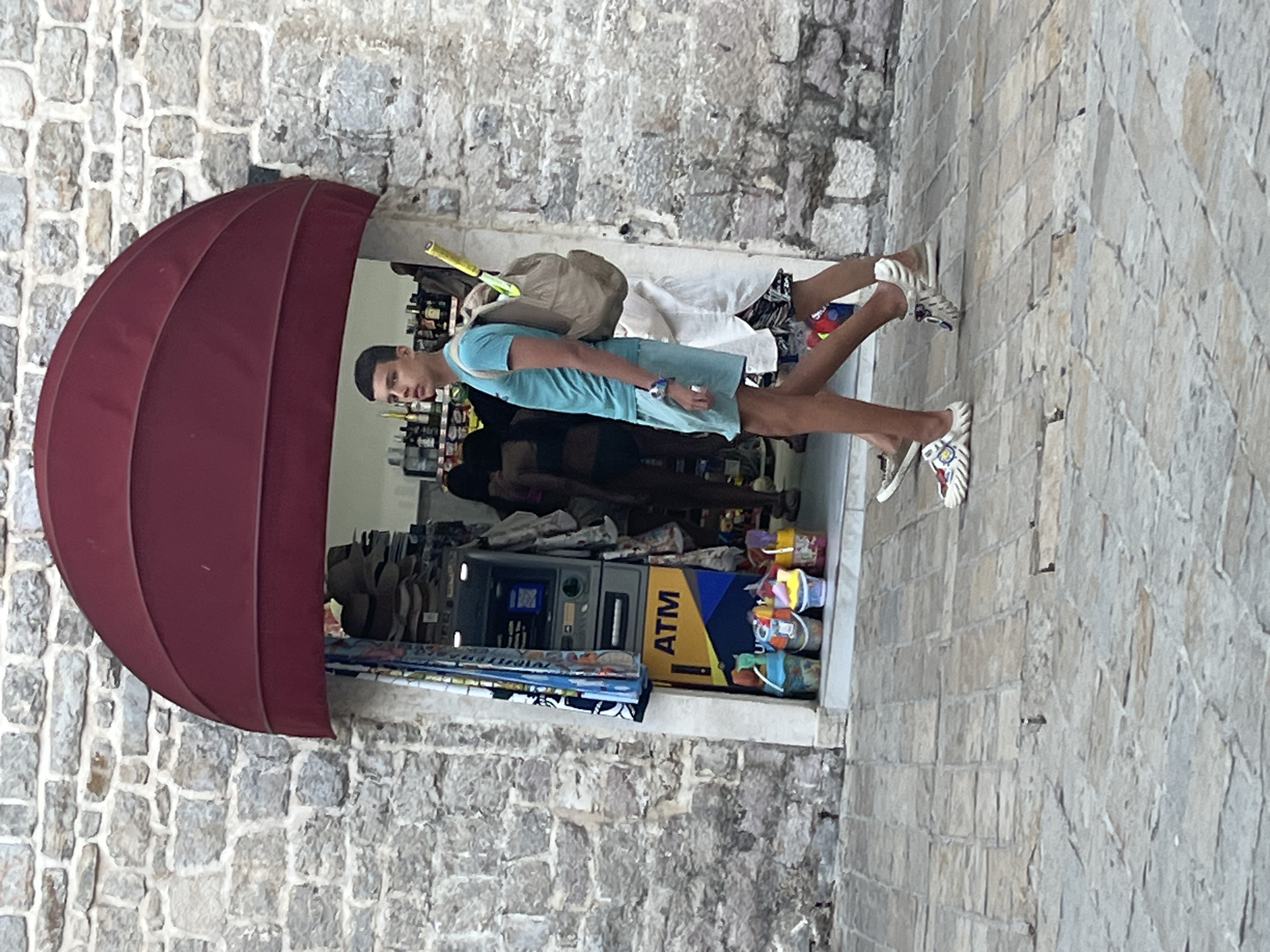 A young man in a teal t-shirt and light blue shorts walks past a store entrance in Budva, Montenegro. He carries a beige backpack over his right shoulder, with a yellow tennis racket protruding from it. Beside him, another person in white shorts and a black and white patterned shirt walks. The store entrance is recessed into a stone building, covered by a burgundy awning. Inside the store, an ATM machine is visible, along with shelves stocked with merchandise and colorful buckets of beach toys. The ground is paved with light-colored stones.