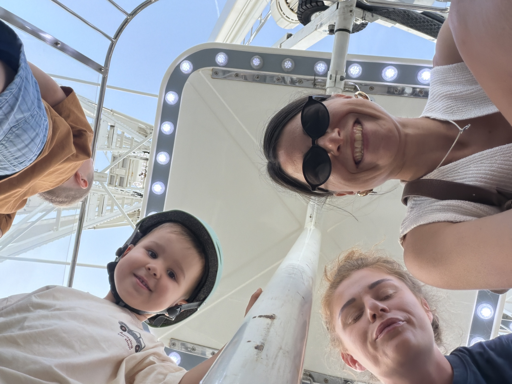 The image captures a vibrant, low-angle perspective of four individuals enjoying a ride on a Ferris wheel, likely in Budva, Montenegro, given the provided context. The bright blue sky, indicative of a clear sunny day, forms the backdrop.
In the top center, a woman with dark hair pulled back wears round black sunglasses and a white textured top, smiling broadly with her teeth showing. A silver necklace is visible around her neck, and a brown strap from a bag is seen over her shoulder. To her upper left, part of a man's head and shoulder are visible, wearing a denim-style shirt over a brown one.
On the bottom left, a young child, possibly a boy, with light brown hair wears a teal helmet with a black strap. He looks upwards with a gentle smile. His cream-colored t-shirt features the visible text "Amazing car show" alongside a small car graphic.
On the bottom right, another woman with light brown, wavy hair looks up, her eyes softly closed in what appears to be a moment of enjoyment or contemplation, a subtle smile on her lips. She wears a dark t-shirt.
A prominent white, slightly weathered pole or support structure runs vertically through the center of the frame. Surrounding the individuals and extending towards the edges are the white interior structures of the Ferris wheel cabin, adorned with rows of glowing, round LED lights embedded in a gray strip. Above, parts of the Ferris wheel's mechanical framework, including spokes and a tire-like component, are visible against the expansive blue sky. The overall mood is one of lighthearted fun and shared experience.