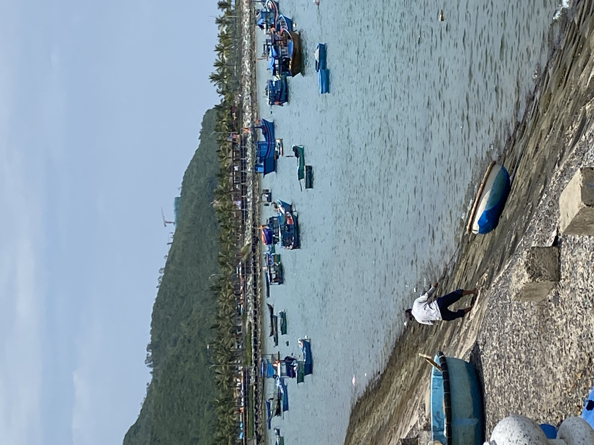 This is a wide-angle, high-angle shot of a bustling fishing harbor in Nha Trang, Vietnam. In the foreground, a man in a white shirt and dark pants is pulling a small blue boat up a sloped, rocky embankment. To his left, a larger blue boat is pulled up on the shore. The water is filled with numerous fishing boats of various sizes, mostly painted blue, anchored in the bay. In the background, a lush green mountain slopes down towards the coast, dotted with palm trees and buildings. The sky is partly cloudy. The image captures a moment of daily life in a Vietnamese fishing community.