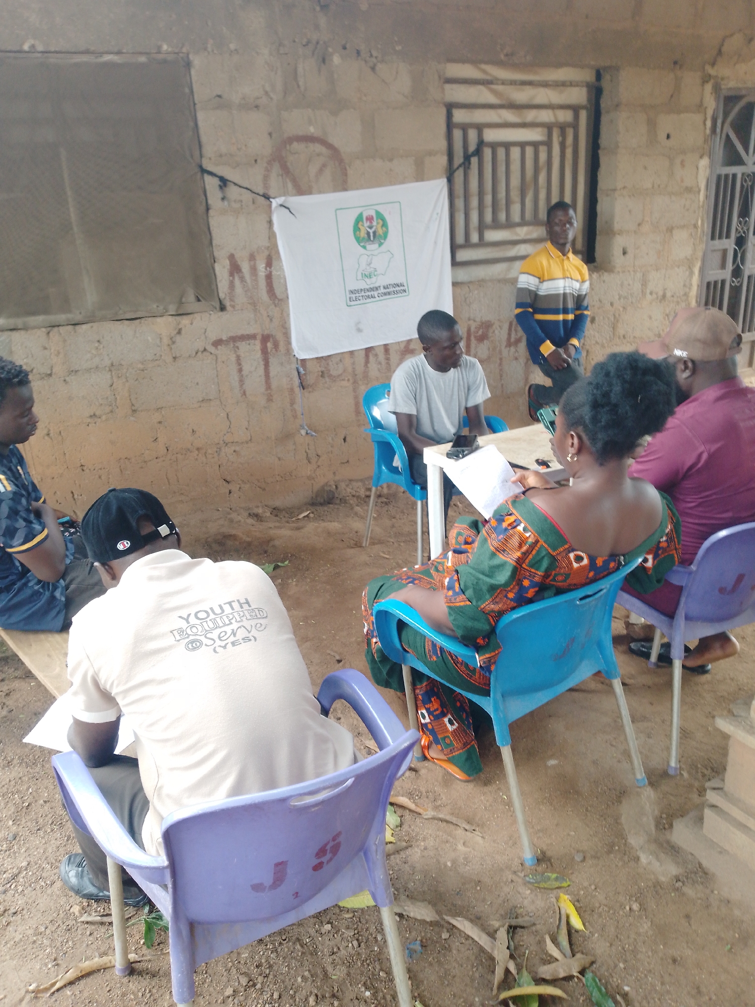 In an outdoor setting with a rough, concrete-block wall as the background, a group of individuals appears to be engaged in a meeting or registration process. A white banner with the emblem of the Independent National Electoral Commission (INEC) is displayed on the wall. In the foreground, two men are seated on a wooden bench, one wearing a light-colored t-shirt with the text "YOUTH EQUIPPED TO SERVE (YES)" on the back. Another man in a grey t-shirt is seated at a white table, looking towards a woman seated in a blue plastic chair. This woman, dressed in a colorful patterned outfit, is holding papers. To her right, a man in a maroon t-shirt and a brown cap is also seated. Further back, a man in a yellow and blue striped shirt stands. The ground is covered in dirt and scattered leaves. The scene suggests an informal administrative or community event.