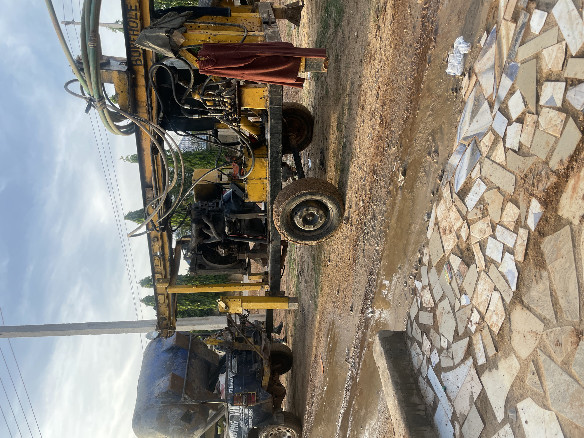A yellow borehole drilling machine is parked on a dirt road in Rido, Nigeria. The machine has a complex system of hoses, cables, and mechanical components. A maroon garment hangs from the side of the machine. A blue water tank is attached to the rear of the machine. The road is muddy, and a section of mosaic tiles are visible on the right side of the road. A utility pole stands on the left side of the machine. The sky is partly cloudy.