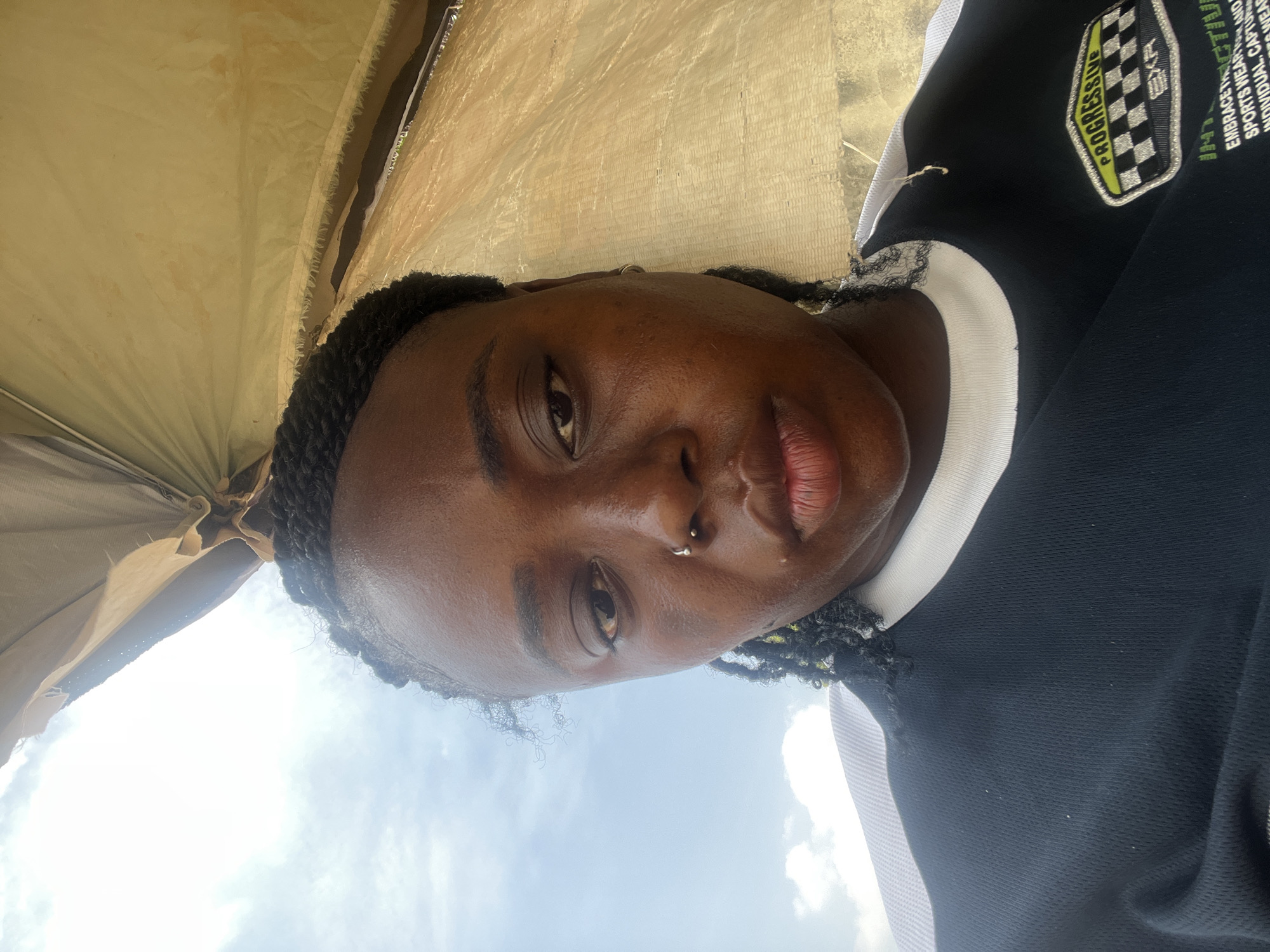 The image shows a close-up, sideways selfie of a young Black woman, with braids framing her face. She is wearing a black jacket with white trim and a patch on the left chest featuring a checkered flag and the words "PROGRESSIVE" and "EXF".
The woman is looking towards the camera with a neutral expression. A silver nose ring is visible. The background is outdoors, with a blurred, yellowish fabric overhead, suggesting she is under a shelter or tent. Sunlight is prominent, creating a bright, possibly midday or late morning scene, with a clear blue sky with some clouds visible through gaps in the fabric. The overall impression is casual and likely taken on a sunny day. The provided location context is Abuja, Nigeria.