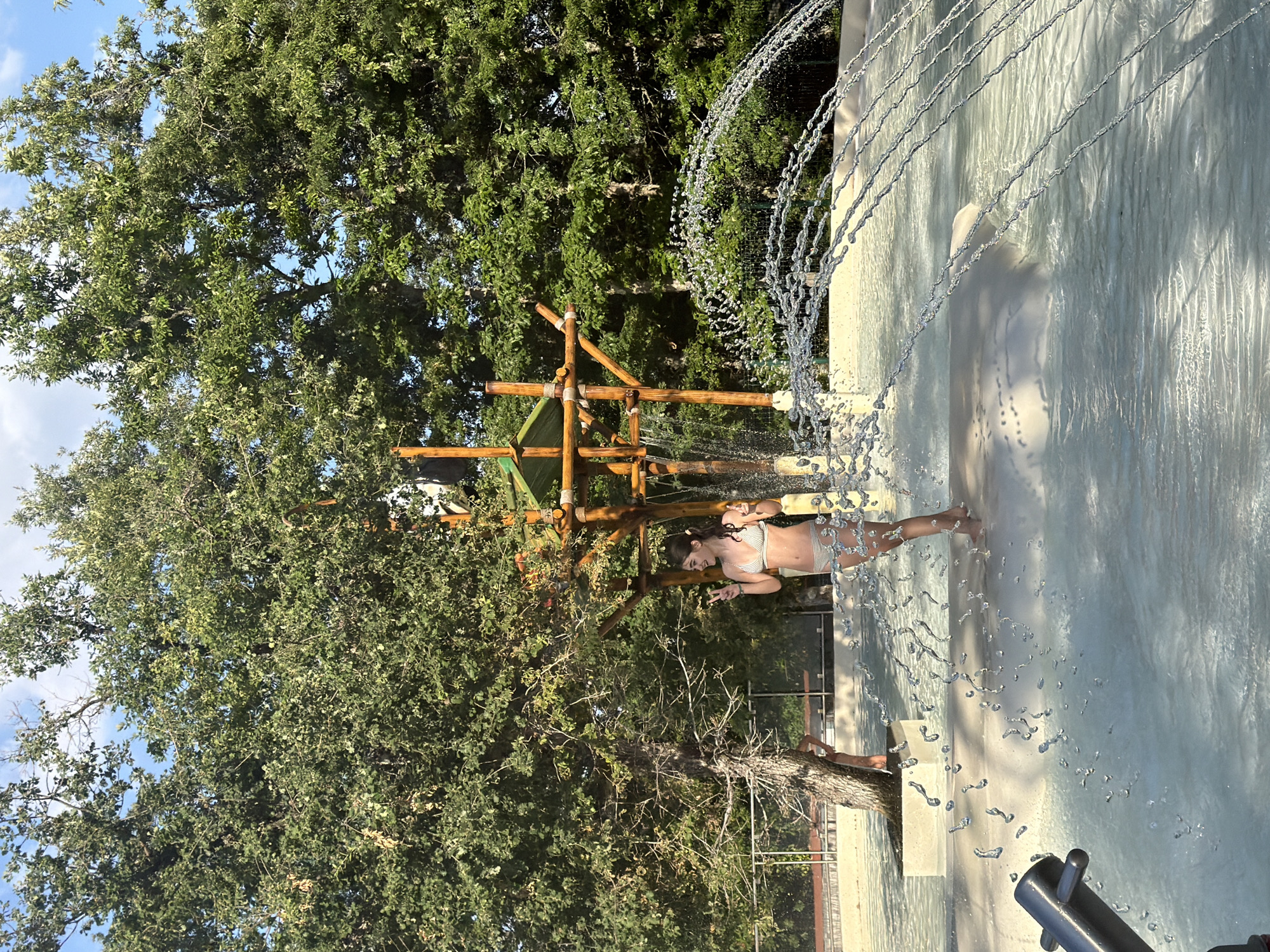 A young woman in a white bikini is standing in a shallow water park, smiling and making a peace sign with her fingers. She is partially obscured by water spraying from a fountain in the foreground. Behind her, a wooden structure with a green roof stands, part of the water park's amenities. The background consists of lush green trees and a blue sky with some clouds. The scene appears to be at the Cavriglia Water Park in Cavriglia, Italy.
