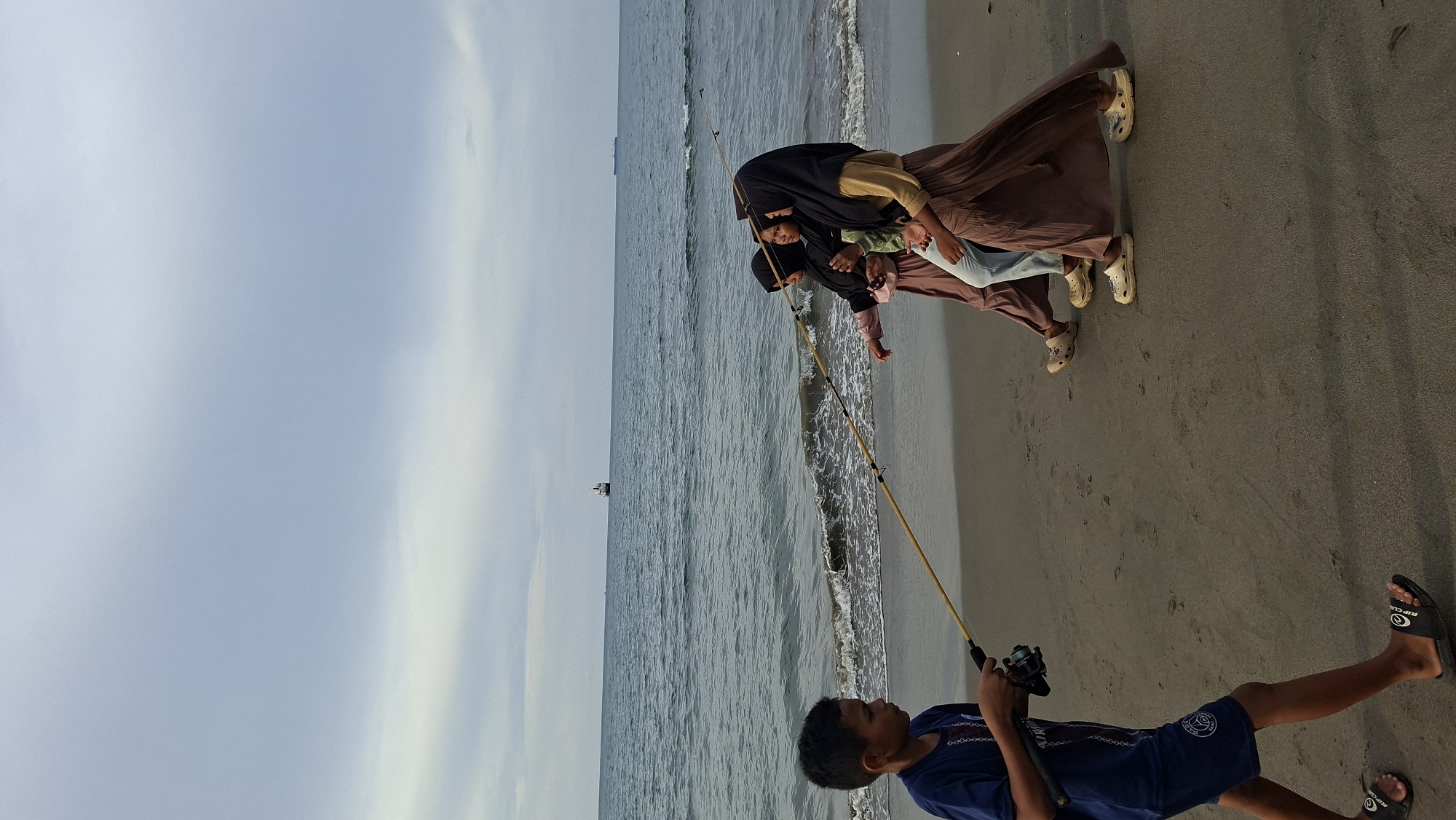 In Lhokseumawe, Indonesia, on a sandy beach, a young boy in a blue t-shirt and shorts is fishing with a rod and reel. He is walking on the beach. Three girls, dressed in hijabs and long skirts, are walking on the beach away from the boy. The sea is calm with small waves, and a large ship is visible on the horizon. The sky is overcast.