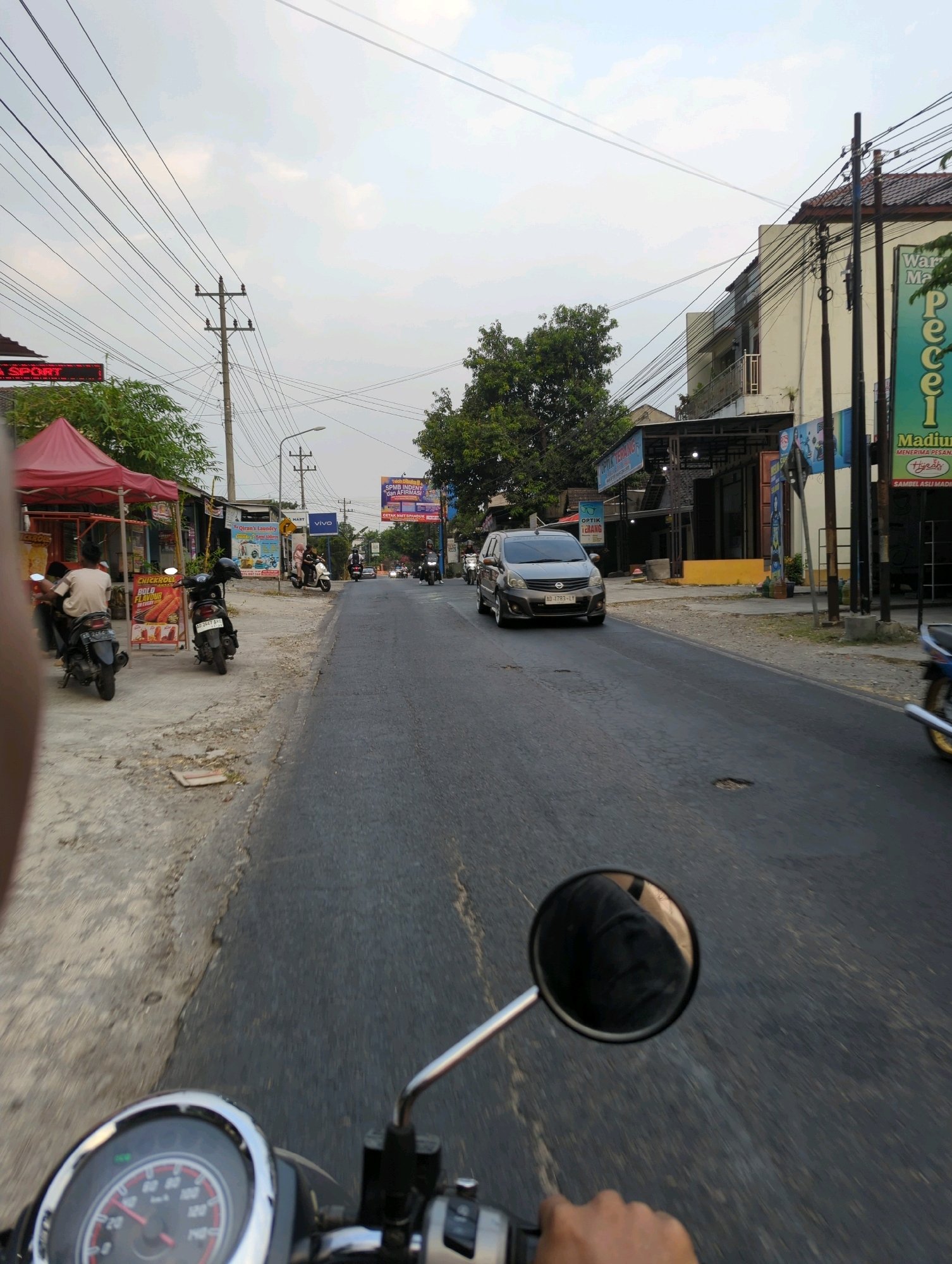 This is a point-of-view photograph taken from a motorcycle, looking forward along a paved road in what appears to be a semi-urban or suburban area. The sky is overcast, suggesting a cloudy day. On the left side of the road, there are several businesses with red and blue awnings. One sign reads "SPORT" in red neon letters. There are also several parked motorcycles and a red tent structure. Further down the left side of the road, a sign for "OPTIK TERANG" is visible.
On the right side of the road, there are buildings, including one with a sign that says "Warung Pecel Madiun." Several telephone poles with numerous wires stretch across the scene. A dark-colored minivan is traveling in the opposite lane, and other motorcycles are also present on the road. The road surface is asphalt and shows signs of wear, with some potholes. In the foreground, part of the motorcycle's handlebars and speedometer are visible, indicating the perspective of the rider. The overall impression is of a busy street scene in a small Indonesian town, likely during daylight hours.
