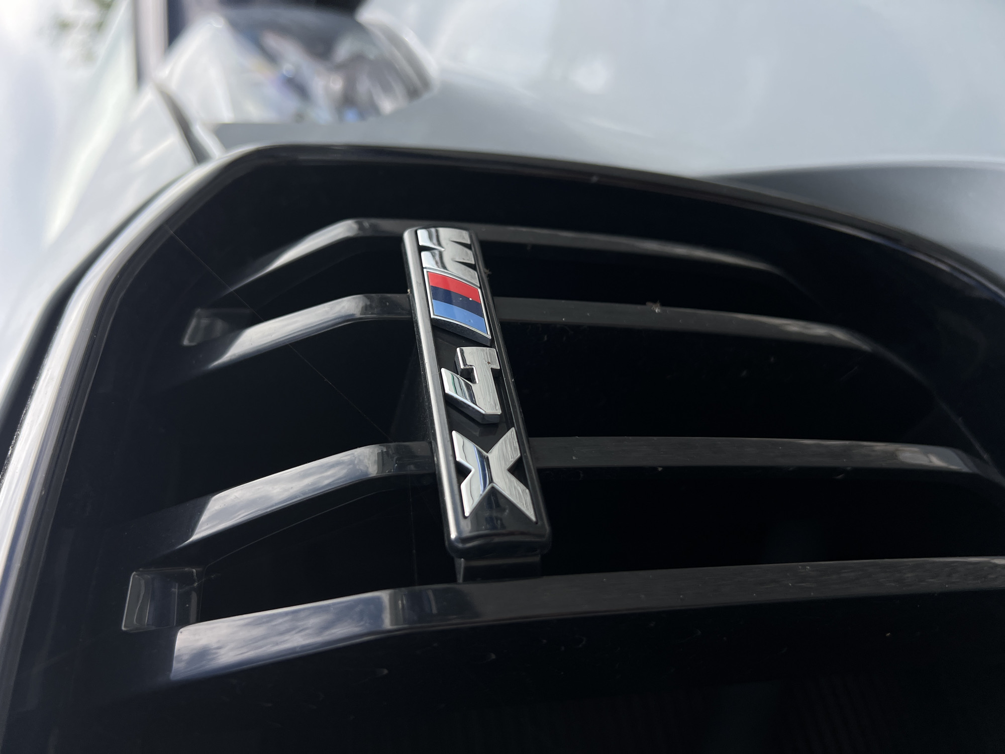 A close-up, shallow depth of field shot focuses on the "X4 M" badge, which is mounted on the black grille of a BMW X4 M. The badge is silver with red, white, and blue M stripes. The car is light-colored, possibly white or silver, and partially visible on the right side of the frame. The background is blurred and appears to be outdoors, with a cloudy sky. The scene is consistent with a vehicle parked on a street or in a parking lot in Cluj-Napoca, Romania. No people or other significant objects are visible in the frame.