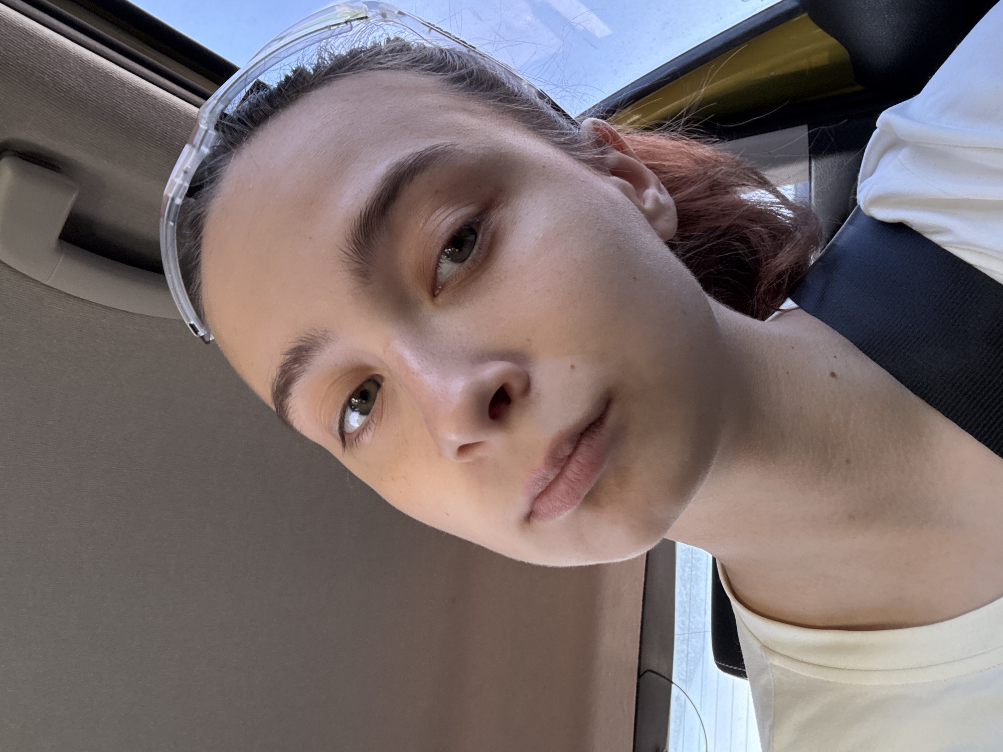 A young person with light skin, wearing transparent safety goggles atop their head, is positioned inside a vehicle. The person has brown hair with red highlights in the back, and is wearing a white t-shirt with a black seatbelt across their chest. The interior of the vehicle is primarily gray and yellow, with a view of the sky visible through the open roof. The location is unconfirmed. The city cannot be determined from this picture.