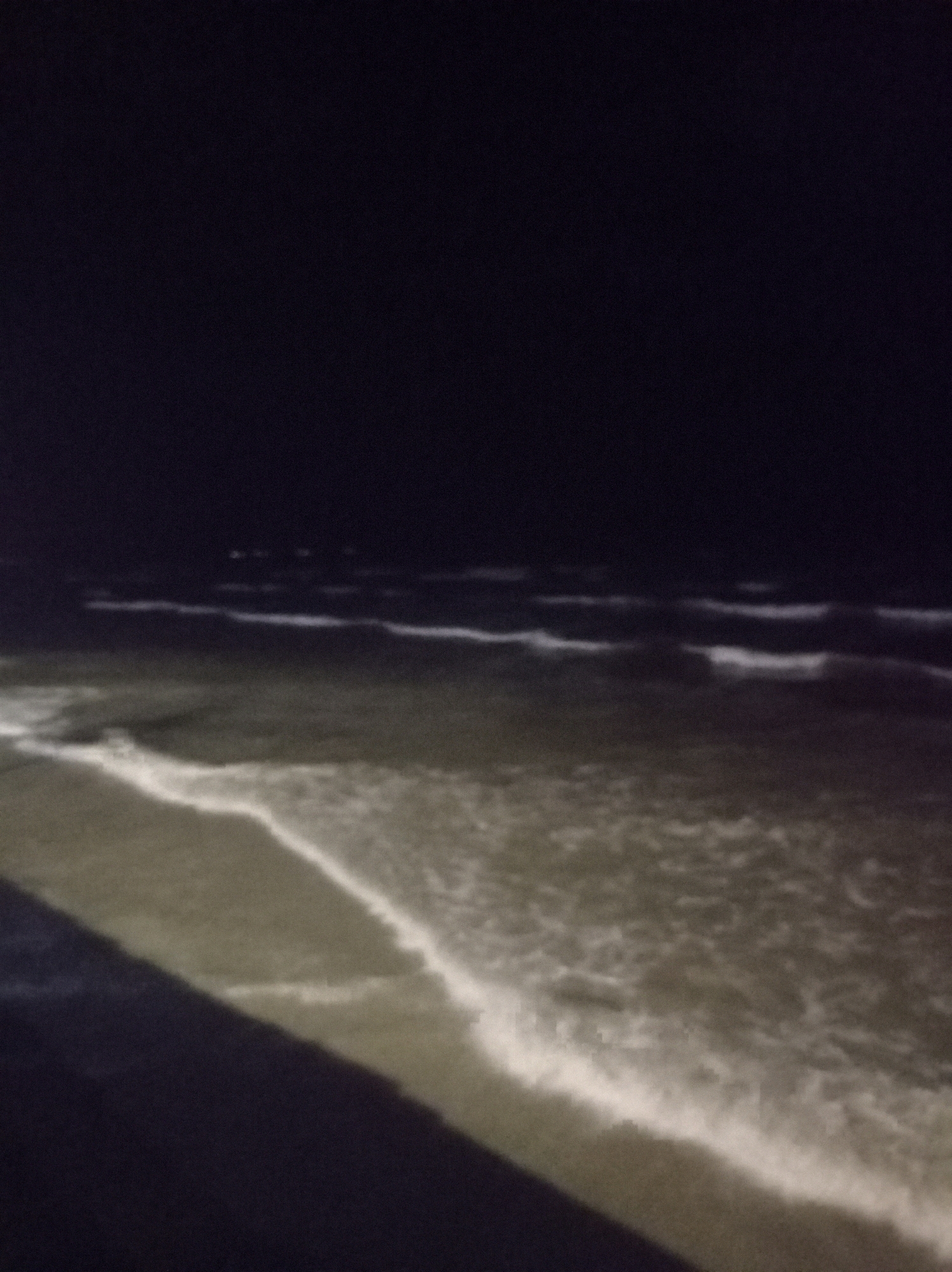 The image shows a beach at night. The sky is dark and there are waves on the ocean. The sand is visible in the foreground. The location is not discernible from the image itself.
