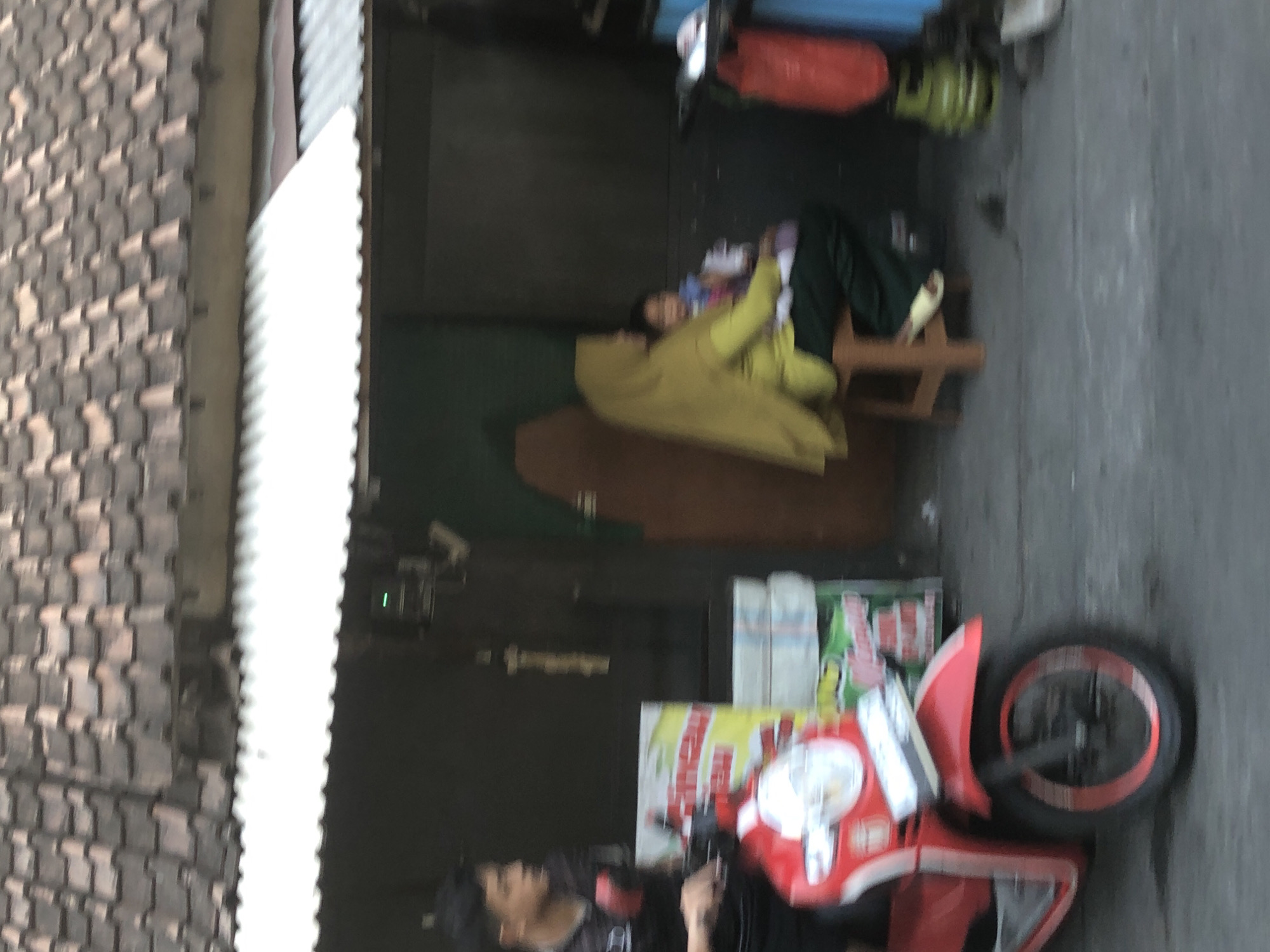 In Tenggulunan, Indonesia, a man wearing a dark t-shirt and dark pants is seated on a red and white motorcycle. He is looking to the left. To his right, a woman wearing a yellow hijab and green pants is seated on a wooden stool, holding a child. The woman is looking forward. Behind them is a dark building with a corrugated metal roof. A red bag and a green gas cylinder are visible to the right of the building. The ground is paved.