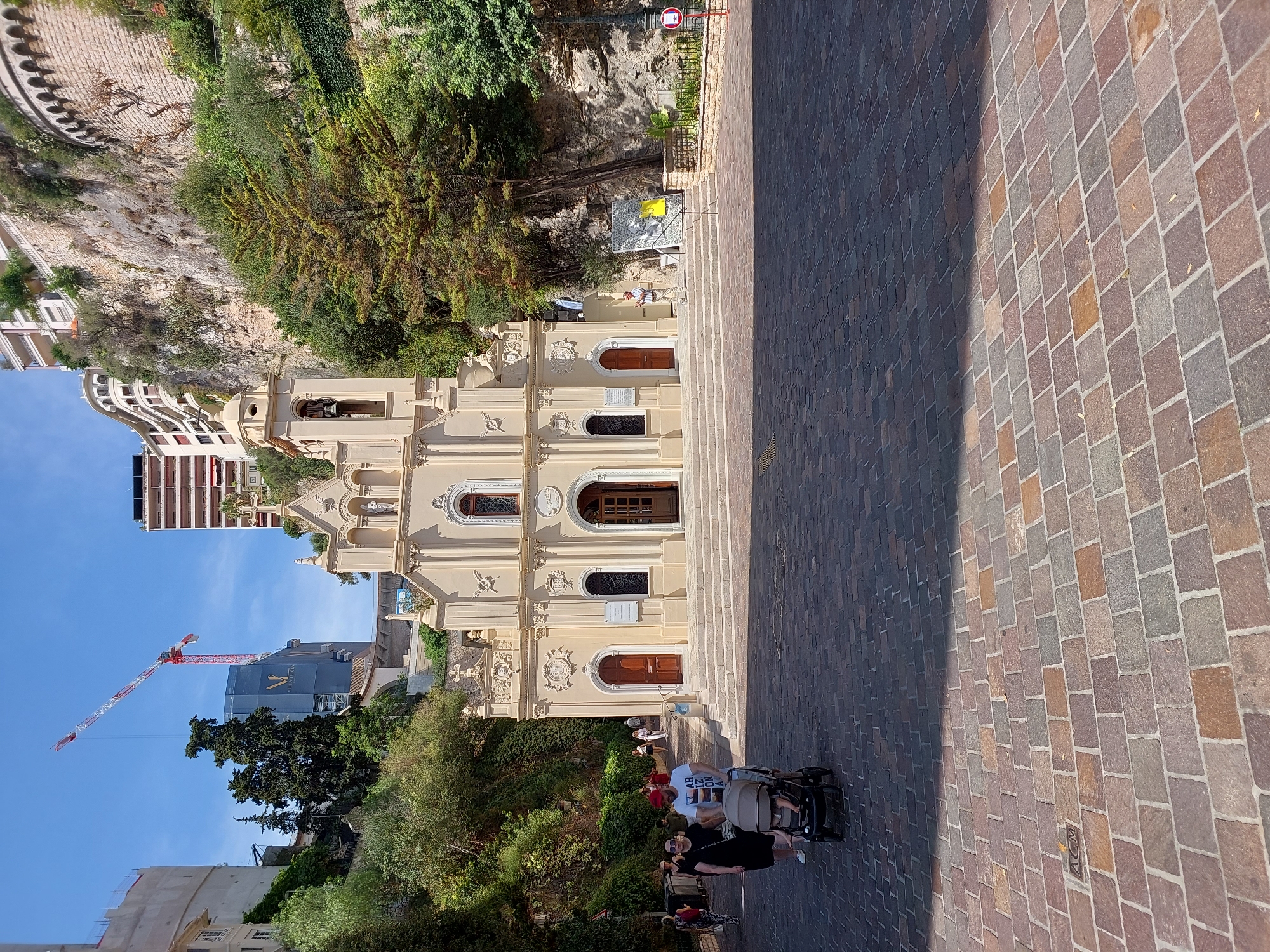 The image shows the Church of Saint Charles and Saint Dévote in Monaco-Ville, Monaco. The church is a pale yellow building with a bell tower and a facade adorned with statues and decorative elements. It is situated on a stone-paved plaza, with a set of stairs leading up to its entrance.
In the foreground, a family is walking across the plaza. A woman in a black dress and sunglasses pushes a stroller with a baby inside. A man wearing a white t-shirt with red lettering and a red cap walks beside them.
In the background, a large construction crane is visible, along with several modern buildings constructed into the side of a cliff. Greenery, including trees and vines, covers parts of the cliff face. The sky is clear and blue. A man in a light-colored shirt and trousers stands on the steps of the church, looking at a mosaic artwork displayed nearby.
