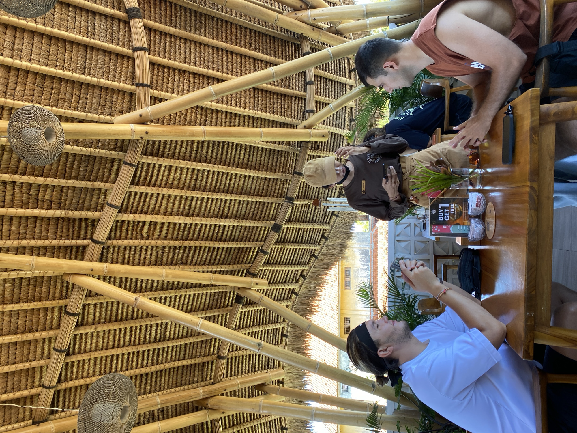 The image shows a scene at a table inside a structure with a thatched roof in Kuta Selatan, Indonesia. Three people are present: two men seated at the table, and one standing to the left. One man, wearing a brown tank top, is seated on the right side of the image and leaning toward the table. To his left, a person in a brown uniform, khaki pants, and a beige hat is seated. A third man, wearing a white t-shirt and a black headband, is seated and turned to the left side of the frame, looking at his hands. A wooden table is present. Items on the table include glasses, a small vase with greenery, and a sign that reads "BUY 1 GET 1 FREE". The structure's roof is made of thatch and supported by bamboo poles. Woven baskets are suspended from the roof as light fixtures or decorative elements. In the background, outside the structure, there is a building with a tiled roof.