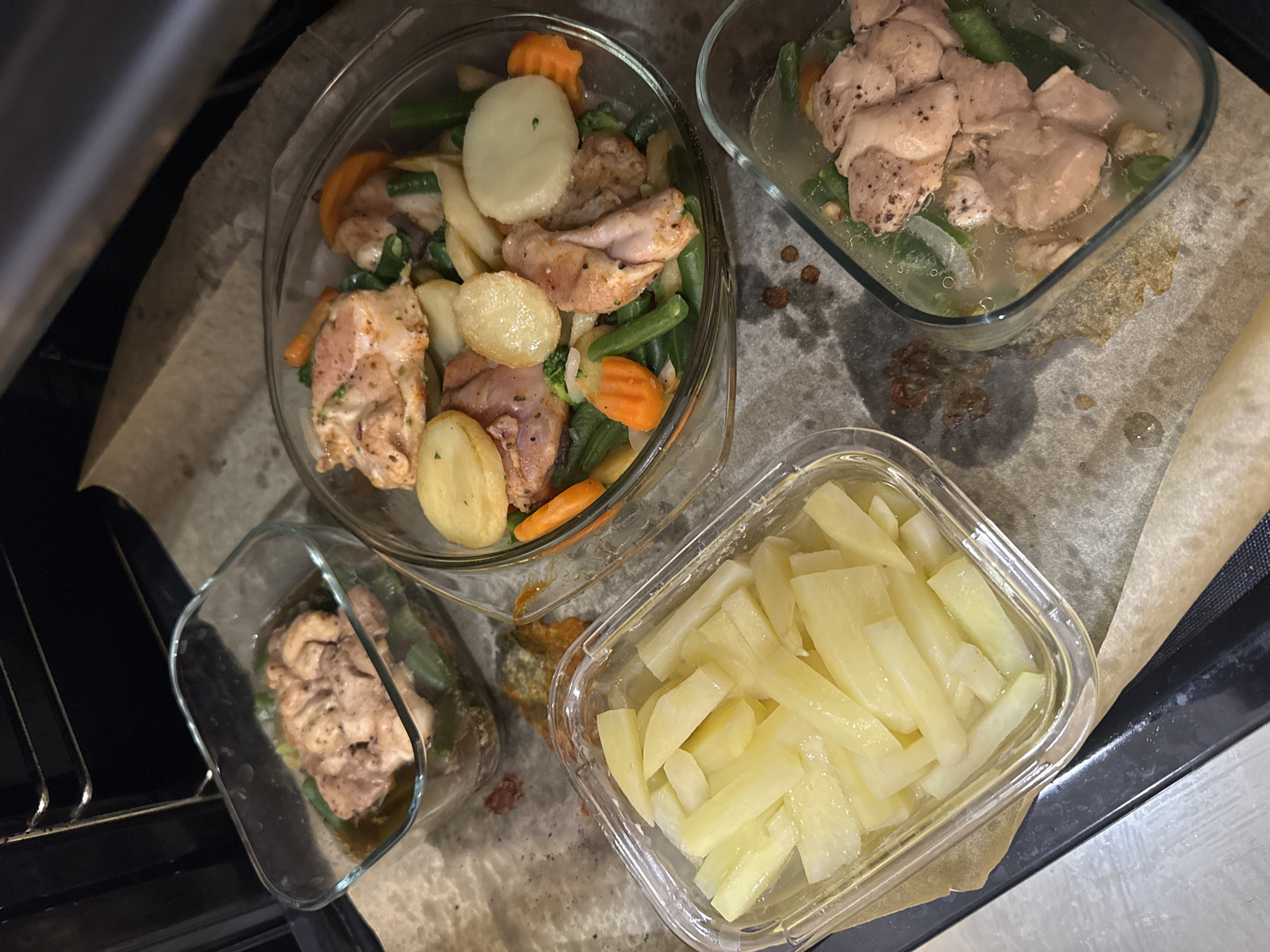 An overhead shot captures four glass containers filled with food, arranged on a sheet of baking paper, seemingly placed near an open oven in a kitchen in Kryvyi Rih, Ukraine.
The central and largest glass bowl contains a mix of diced chicken pieces, sliced and small whole potatoes, green beans, and round-cut carrots. The chicken has a light seasoning, suggesting it is either marinated or partially cooked.
In the upper right, a rectangular glass container holds chunks of chicken and some green vegetables submerged in a clear liquid, possibly broth or marinade. A similar smaller container is visible in the bottom left, partially obscured by the dark interior of an oven door or rack, also containing chicken and green vegetables in liquid.
The fourth container, situated in the bottom right, is a clear rectangular dish filled with uniformly cut potato sticks, likely raw or par-boiled, also submerged in a clear liquid.
The baking paper beneath the containers shows some liquid and food remnants, indicating an ongoing food preparation or cooking process. The dark, metal structure on the left and bottom edges of the frame is consistent with an open oven or its rack. The overall scene suggests a meal, possibly chicken and vegetable bake, being prepared for cooking. There are no people or visible text in the image.