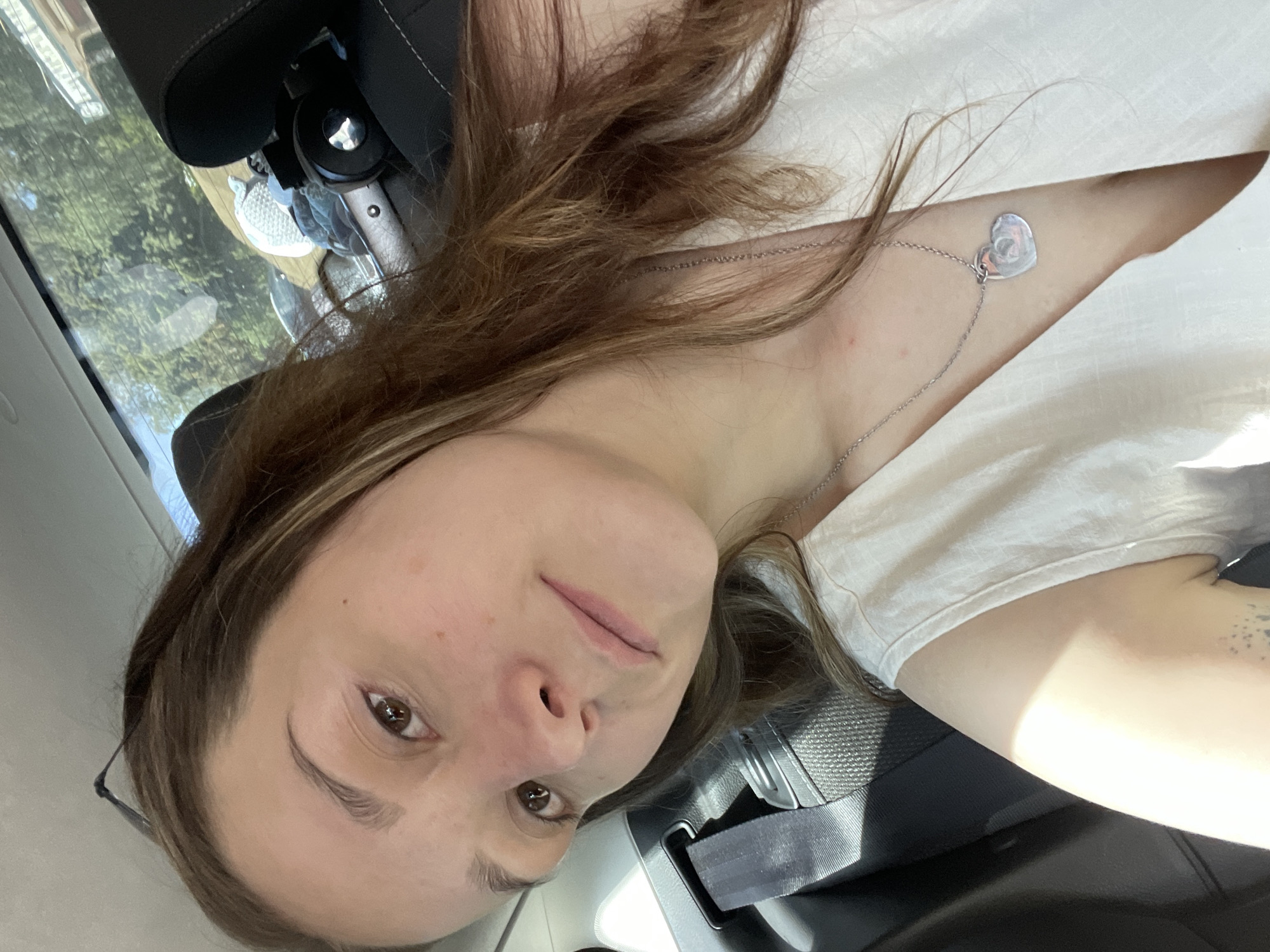 A young woman with long, brown hair is taking a selfie in a car. She is wearing a white t-shirt and a silver necklace with a heart-shaped pendant. Her face is partially visible, and she is looking at the camera with a neutral expression. The background shows the interior of the car, including the headrest of a seat and a seatbelt. Sunlight is streaming in through the window, creating shadows on the woman's arm and clothing. The scene appears to be taking place during the daytime, likely in a warm climate, given the sunlight and the woman's attire. The overall impression is a casual, personal moment captured on a bright day.
