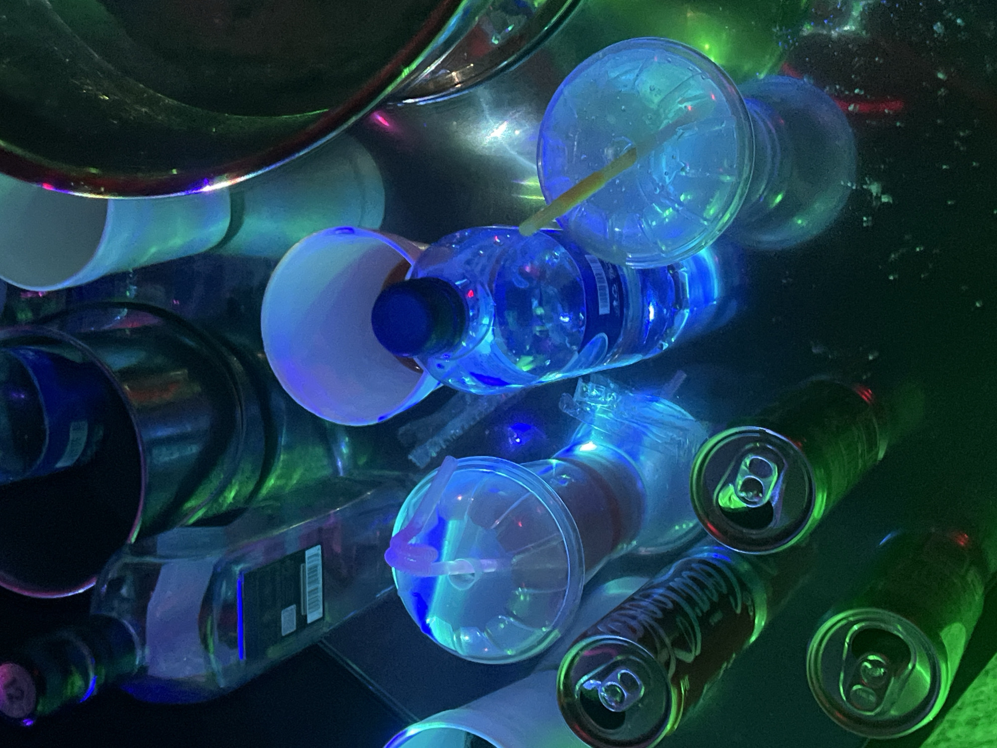 The image shows a collection of discarded items on a reflective surface. The items include beverage cans, plastic cups with lids and straws, a water bottle, a liquor bottle, and white paper cups. The objects are illuminated by blue and green light, which creates a neon-like effect. There is no indication that this image was taken in Jalingo, Nigeria.