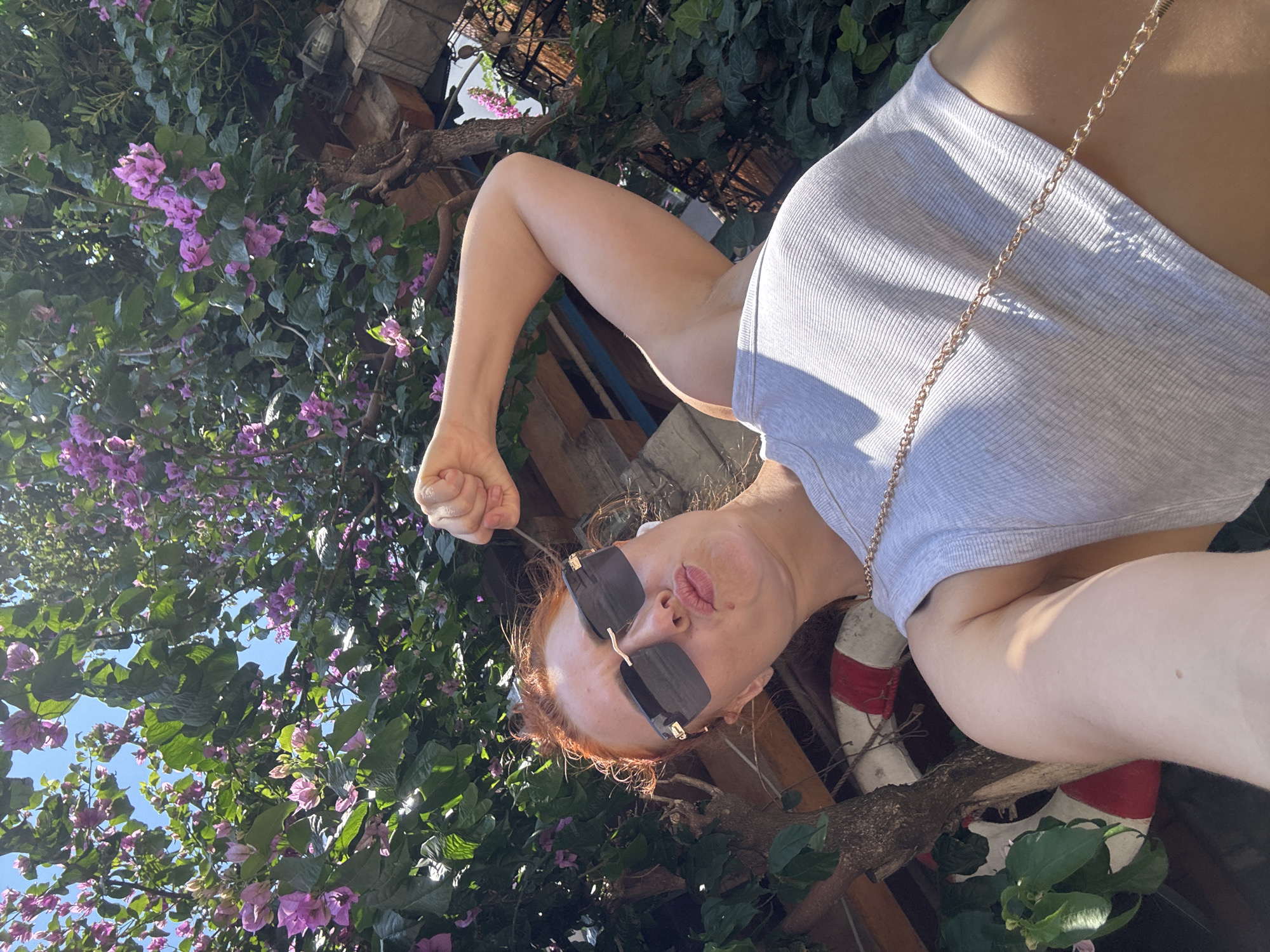 In Budva, Montenegro, a woman with red hair is taking a selfie outdoors. She is wearing sunglasses and a grey crop top, and she is flexing her bicep. The background is filled with greenery and blooming pink flowers. The woman is positioned in the foreground, looking towards the camera with her lips pursed.