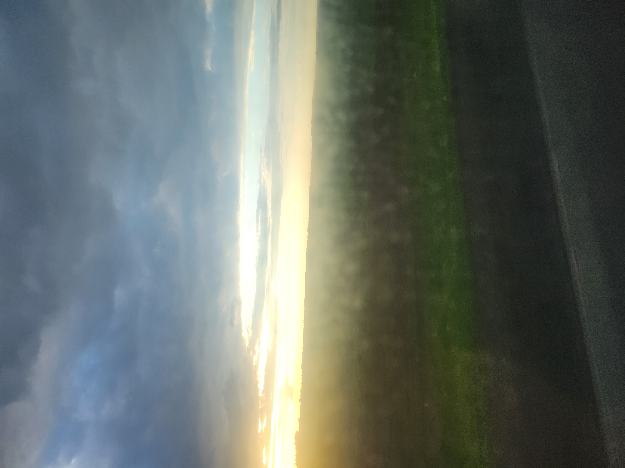 The image depicts a landscape at either sunrise or sunset. On the left side, there is a cloudy sky with shades of blue and gray. Towards the center, there is a bright band of golden and yellow light, suggesting the sun. To the right of the light is a field of green, possibly grass. On the far right, there is a dark, blurred area that could be a road or another dark surface. The location is unknown.