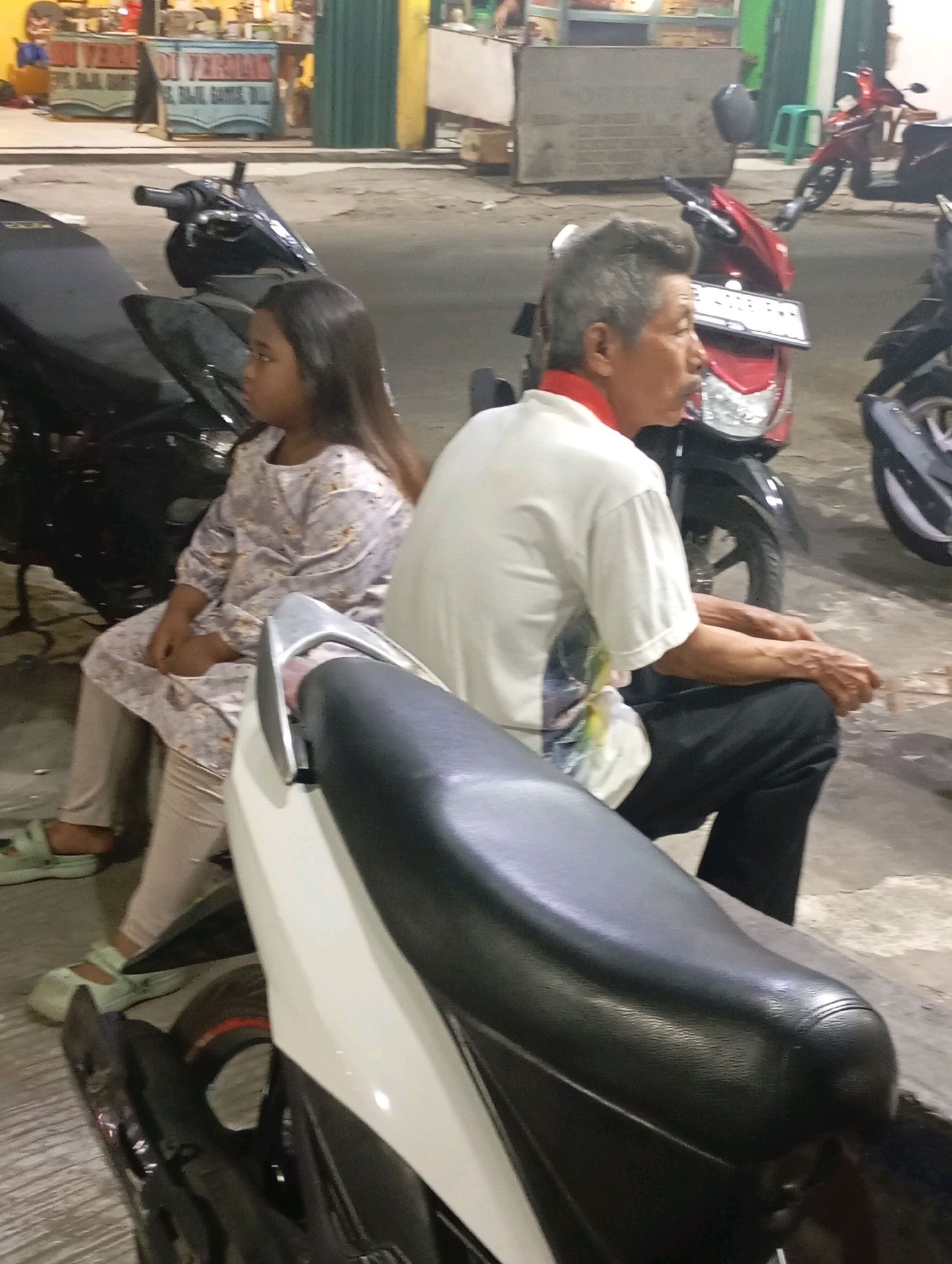 In Babelan, Indonesia, an outdoor scene at night shows two individuals seated on motorcycles parked on a paved surface. A young girl with dark hair is positioned on the left, wearing a light-colored patterned top and beige pants, with light green sandals on her feet. To her right sits an elderly man with gray hair, a mustache, and a red collar on his white shirt, wearing black pants. Both are looking away from the camera, with the man's hands resting on his lap. In the background, a red motorcycle is visible, and further back, food stalls with signage are illuminated. The overall lighting suggests an urban night setting.