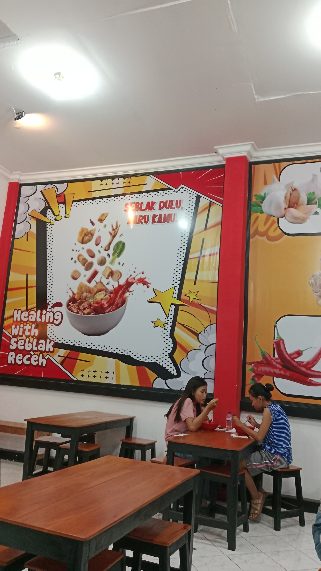 This image shows the interior of a restaurant, likely a warung or cafe, in Blitar, Indonesia. Two young women are seated at a dark-colored wooden table with a reddish-brown top, eating from bowls. One woman on the left wears a pink t-shirt and jeans, while the woman on the right wears a blue and white striped shirt and gray shorts. They are both looking down at their food. A red plastic bottle is visible on the table between them.
The restaurant features colorful murals on the walls. The mural directly behind the women depicts a bowl of food with various ingredients suspended in the air above it, along with text that reads "SEBLAK DULU, BARU KAMU" and "HEALING WITH SEBLAK RECEH". Another mural to the right shows garlic and red chili peppers.
The environment includes several dark-colored wooden tables and matching stools. The floor is tiled with a checkered pattern. The ceiling is white and has some visible lighting fixtures. The overall impression is of a casual dining establishment.
