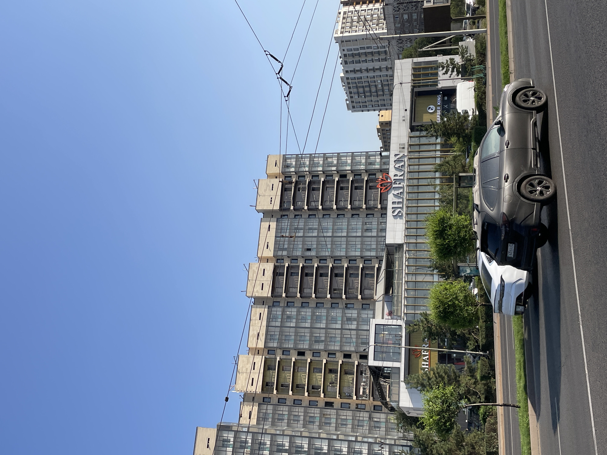 An outdoor, eye-level, medium shot captures a street view in Almaty, Kazakhstan. The scene is dominated by a modern, multi-story residential building with a glass facade and balconies, constructed of light-colored concrete and stone. In front of the residential building, a commercial structure with a glass storefront and a prominent sign reading "SHAFRAN" is visible. To the right, another multi-story building with a white facade and numerous windows stands. The foreground features a paved road with two cars: a white sedan and a dark gray SUV. A narrow strip of grass separates the road from a sidewalk lined with trees. Overhead, power lines crisscross the blue sky. The scene is well-lit, indicating a sunny day.