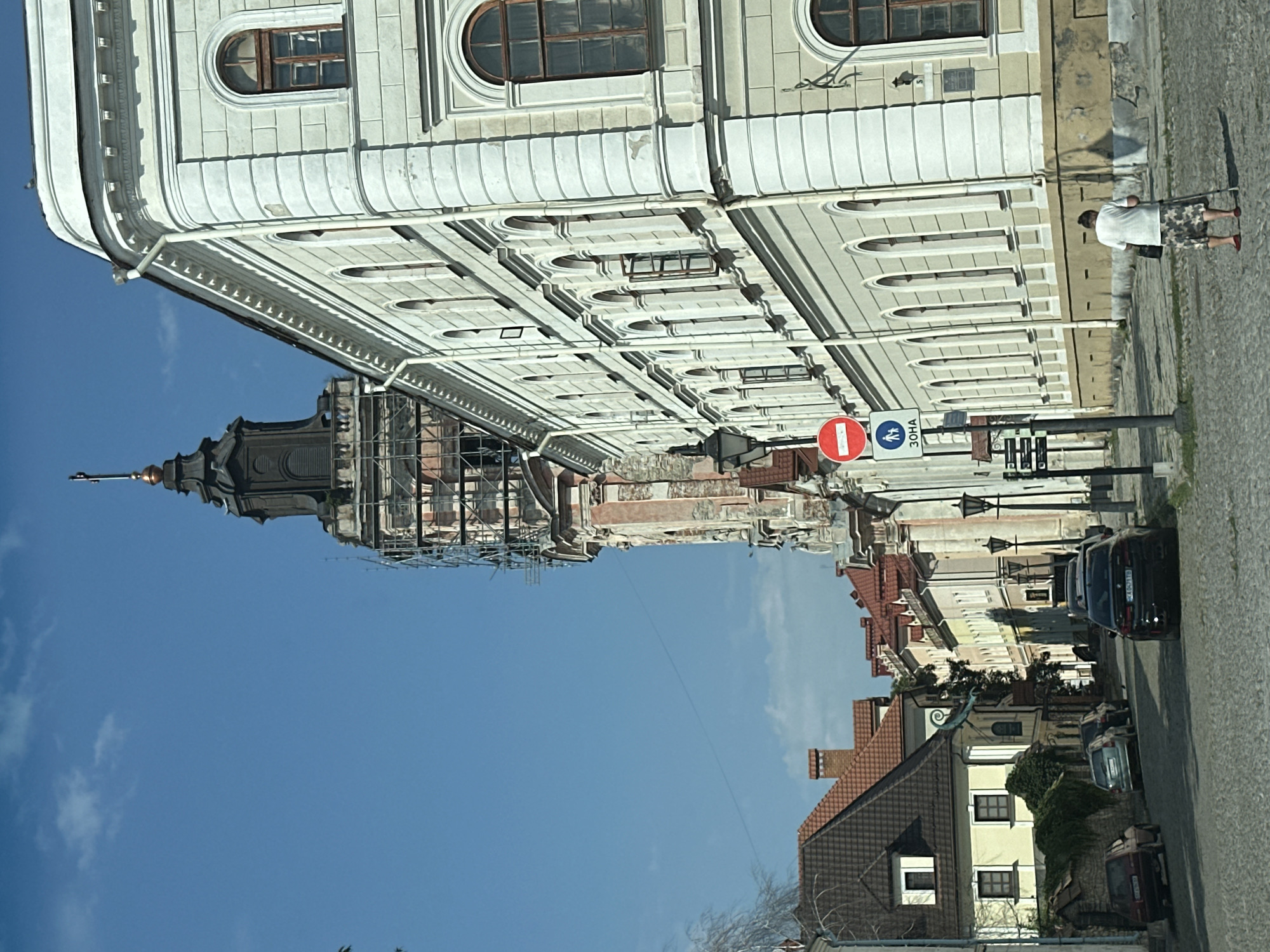 The image depicts a view looking down a cobblestone street in Kamianets-Podilskyi, Ukraine. On the right side of the street, a large, pale yellow building with white trim and numerous arched windows dominates the foreground. Its facade is detailed with decorative elements. Further down the street, other buildings with red tiled roofs are visible.
To the left, a tall, imposing tower is partially obscured by scaffolding, suggesting ongoing renovation or construction. The tower has a dark, multi-tiered roof topped with a cross. Next to the tower, a series of buildings with red roofs and stucco facades line the street.
A person, seen from behind, is walking with the assistance of crutches on the right side of the street, heading away from the camera. They are wearing a white t-shirt and a patterned skirt. Several cars are parked along the street, including a black sedan in the foreground and other vehicles further up the road. Traffic signs are visible, including a circular red sign with a white bar, indicating prohibition, and a square blue sign with white figures of pedestrians. The sky is clear and blue with some scattered clouds. The overall impression is that of an old European town with historical architecture.