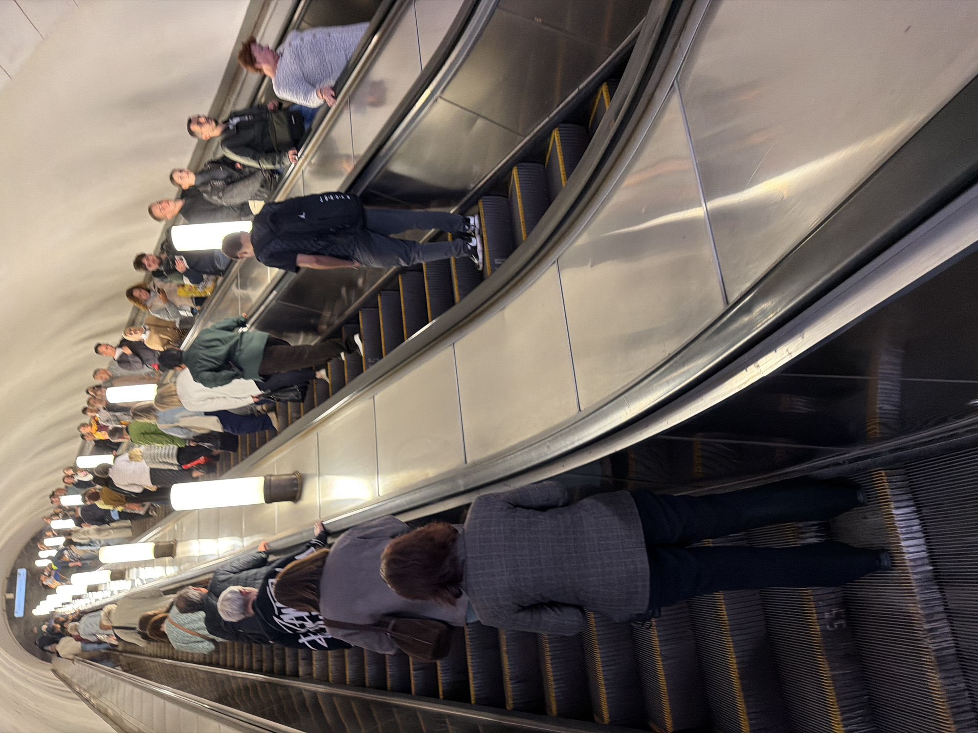 The image shows a wide escalator in what appears to be a subway station, with many people ascending. The escalator is well-lit by overhead lights and several vertical cylindrical lights lining the sides. People of various ages and ethnicities are seen on the escalator, dressed in casual and business attire. Some are carrying bags, and one person is wearing a backpack. The escalator steps are metallic and ribbed, with yellow safety markings. The walls of the station are curved and appear to be made of a light-colored material, possibly concrete or plaster. The scene depicts a busy public space with people commuting.