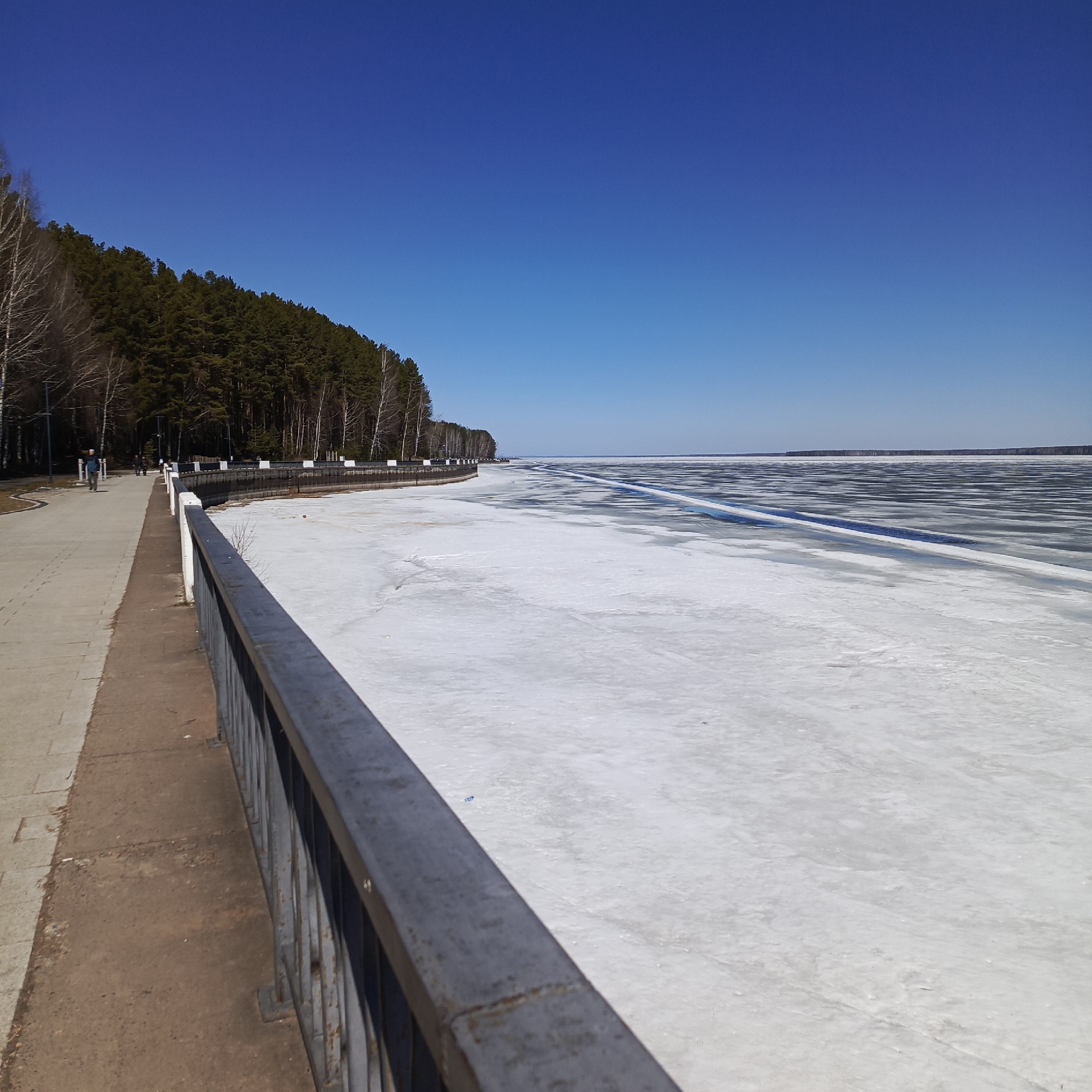 The image shows a partially frozen body of water, possibly a river or lake, with a concrete walkway and metal railing in the foreground. A line of trees is on the left side of the water. The sky is clear and blue. People can be seen walking on the walkway. The scene is in the city of Chaikovsky, Russia. The surface of the water is covered in patches of ice and open water. The rail appears rusted and weather-beaten.