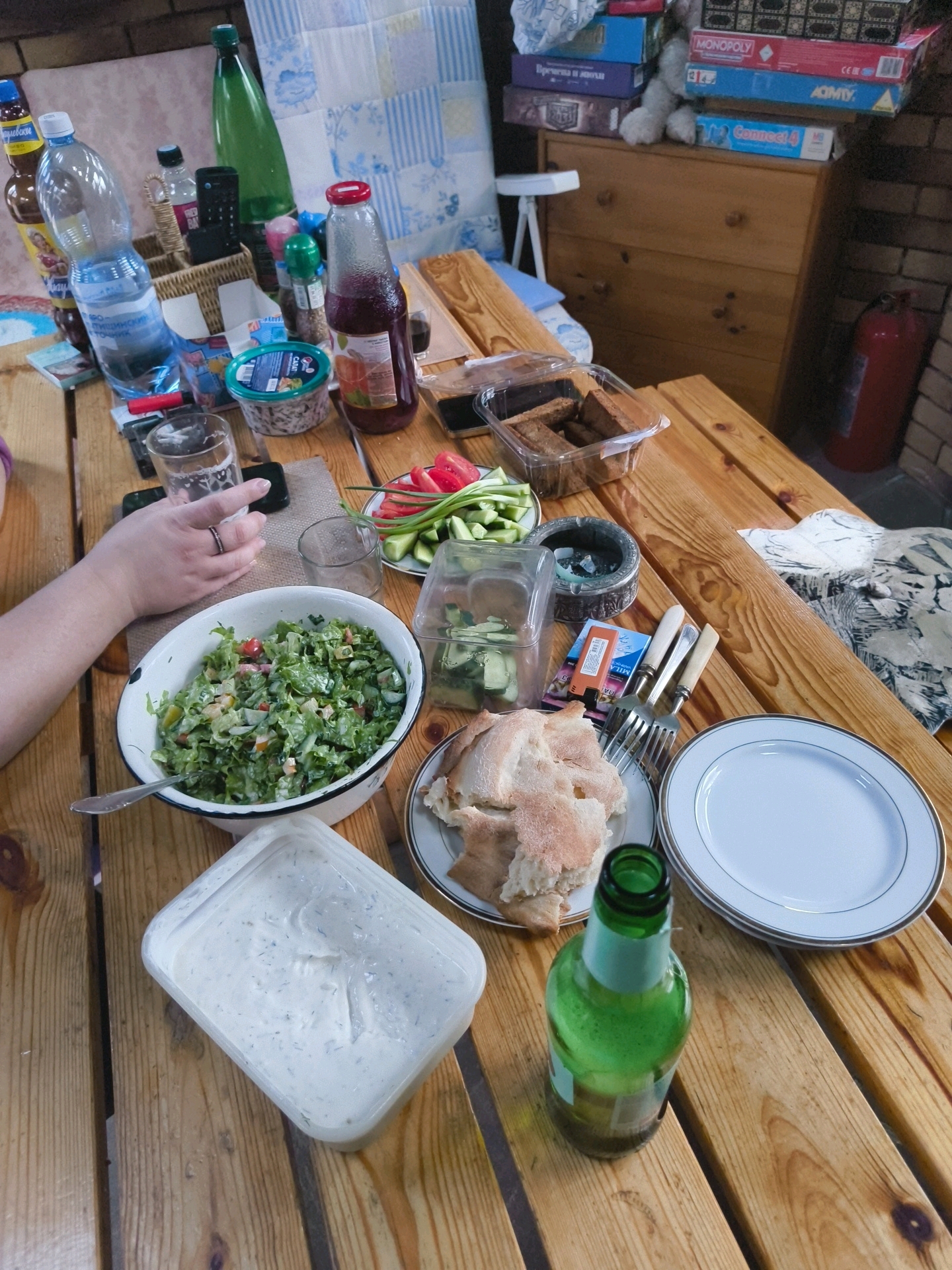 An outdoor wooden picnic table is set with various food items and drinks. In the foreground, a white plastic container filled with a creamy white sauce, possibly dip or dressing, sits on the table. To its right, a white plate holds pieces of flatbread. A green glass bottle, appearing to be a beverage, is positioned next to the bread. Further back, a large white bowl contains a green salad. A plate of sliced vegetables, including tomatoes and cucumbers, is next to the salad. Other items on the table include a plastic container with what looks like breadsticks or small sandwiches, a glass of water, and several bottles of drinks. In the background, a wooden dresser is visible, stacked with board games. A red fire extinguisher is mounted on a brick wall. The scene suggests a casual outdoor meal. The location is specified as Востец Снт, Russia.