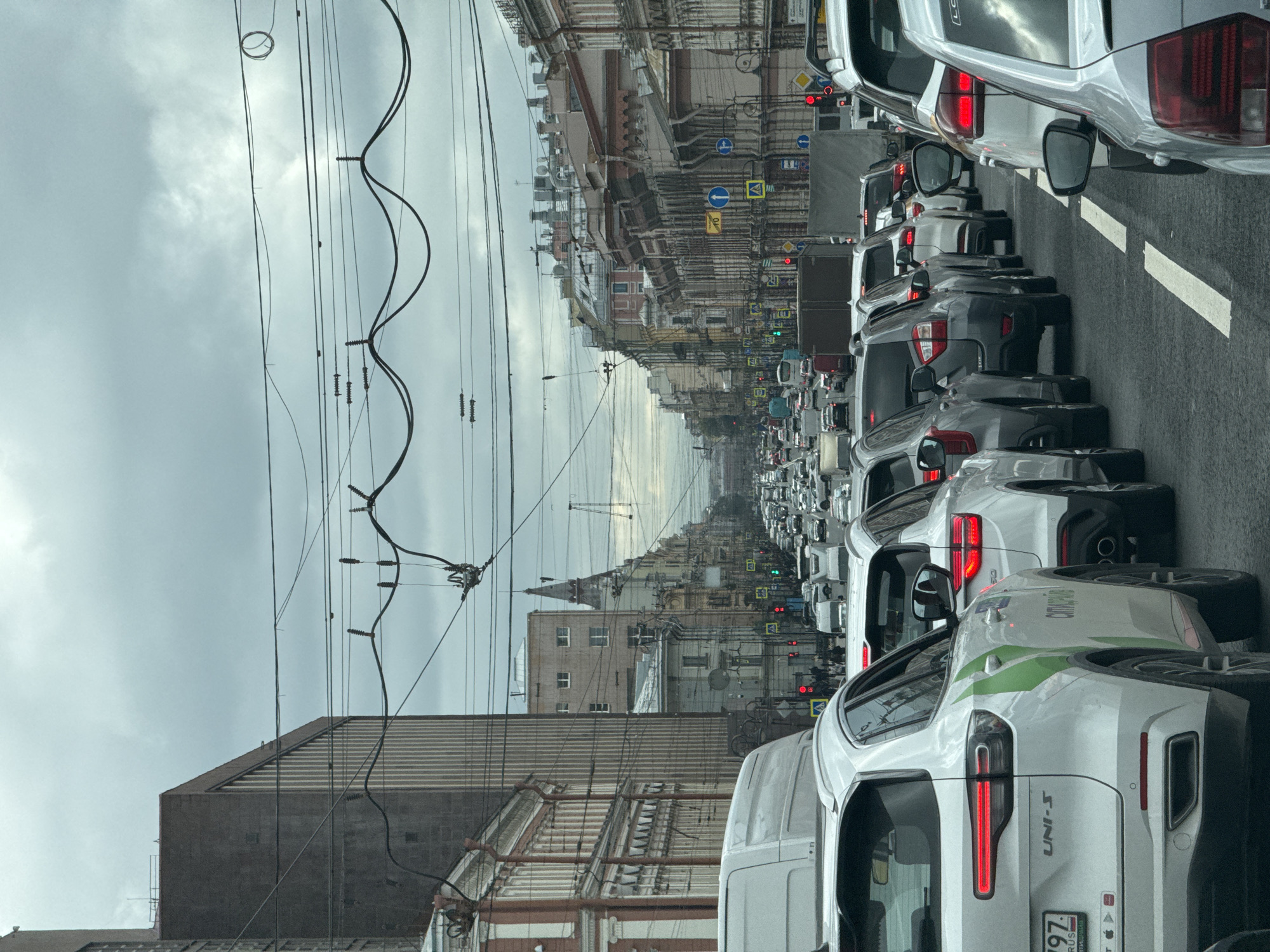 The image depicts a traffic jam on a cloudy day in Saint Petersburg, Russia, likely on Nevsky Prospekt given the architecture and the presence of trams. The scene is dominated by rows of cars, including white SUVs and other vehicles, stuck in traffic. Various buildings line the street, exhibiting classic Saint Petersburg architectural styles with ornate facades and rooftops. Overhead, a complex network of electrical wires and poles crisscrosses the sky. Traffic lights are visible, some showing red, indicating the standstill. Pedestrians are sparsely visible on the sidewalks. The overall impression is one of urban congestion under an overcast sky.