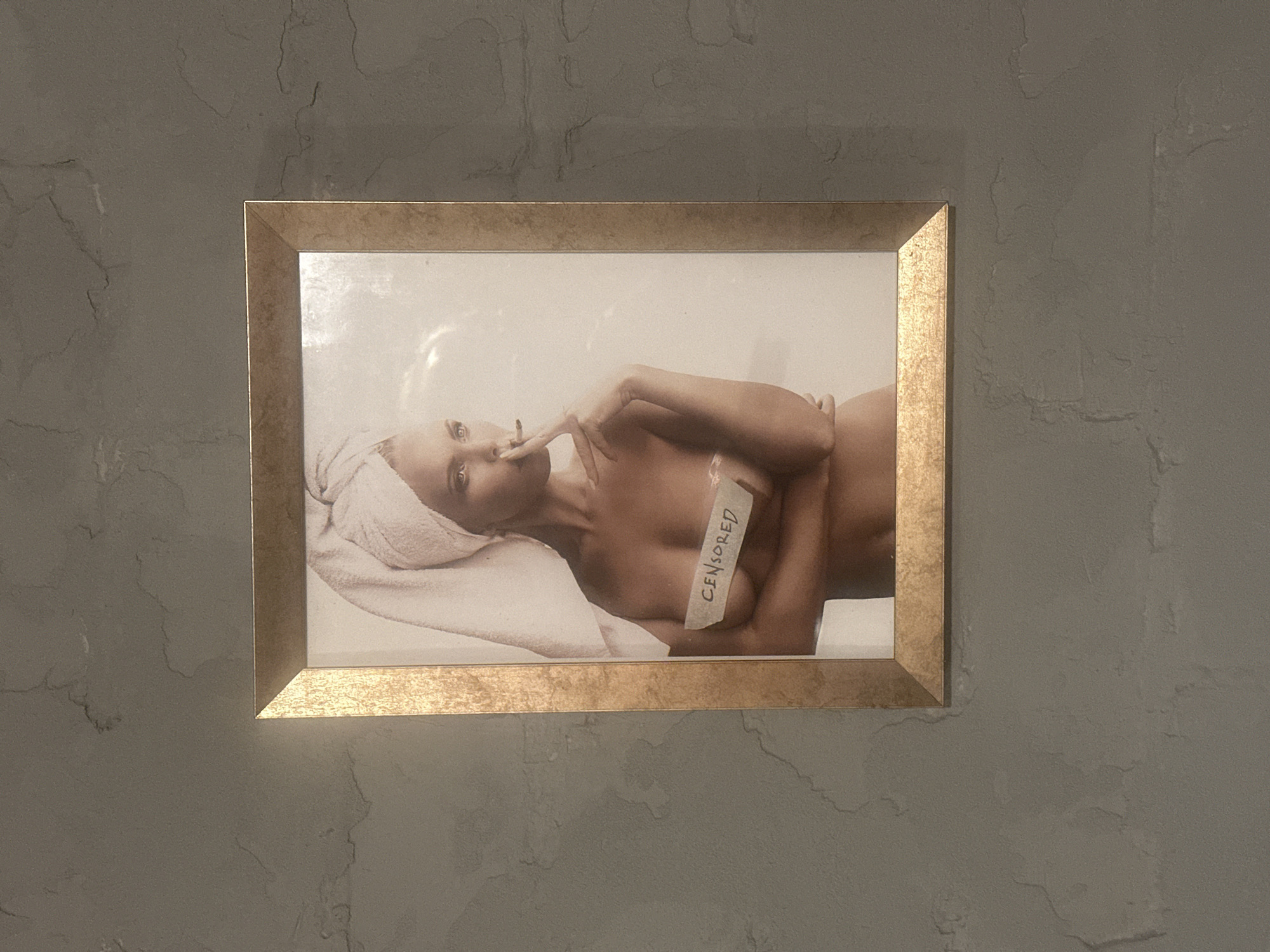 A framed photograph is mounted on a textured gray wall. The photograph features a woman with a towel wrapped around her head, holding a cigarette to her lips. A strip of tape with the word "CENSORED" written on it is placed over her chest. The frame is golden and appears to be made of metal. The wall has a rough, mottled texture with variations in shade. The image and its placement are consistent with decorative artwork in a private residence or establishment in Lapės, Lithuania.