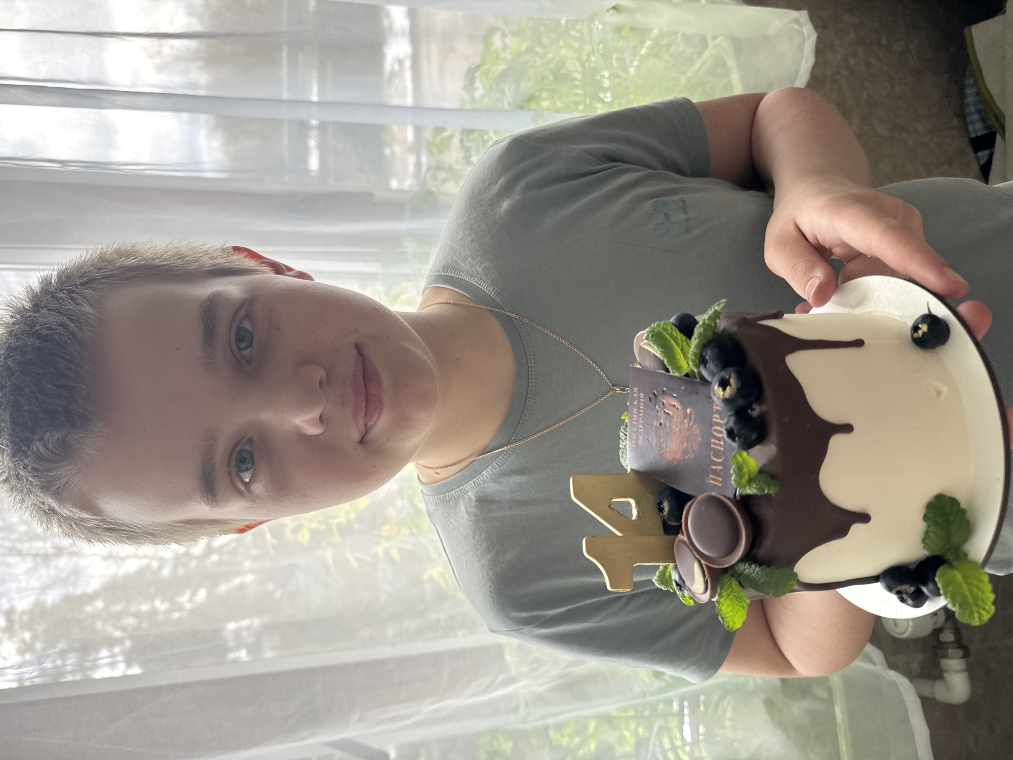 A young person, likely male, with short, light-colored hair and blue eyes, smiles faintly while holding a birthday cake indoors. The image is brightly lit by natural daylight coming through a window covered by sheer white curtains, with blurred green foliage visible outside.
The person is wearing a plain olive-green t-shirt and a thin gold chain. They are holding a round cake decorated with white frosting, dark chocolate drips, fresh blueberries, and mint leaves. A golden number "14" is placed on the cake, indicating a 14th birthday celebration. Also adorning the cake is a miniature replica of a Russian passport, bearing the Cyrillic text "РОССИЙСКАЯ ФЕДЕРАЦИЯ" (Russian Federation) and "ПАСПОРТ" (Passport). This detail suggests the celebration marks the occasion of receiving a passport, which is a significant milestone at age 14 in Russia.
The scene conveys a joyful and personal moment, likely taking place in a home in Раздолье, Russia.
