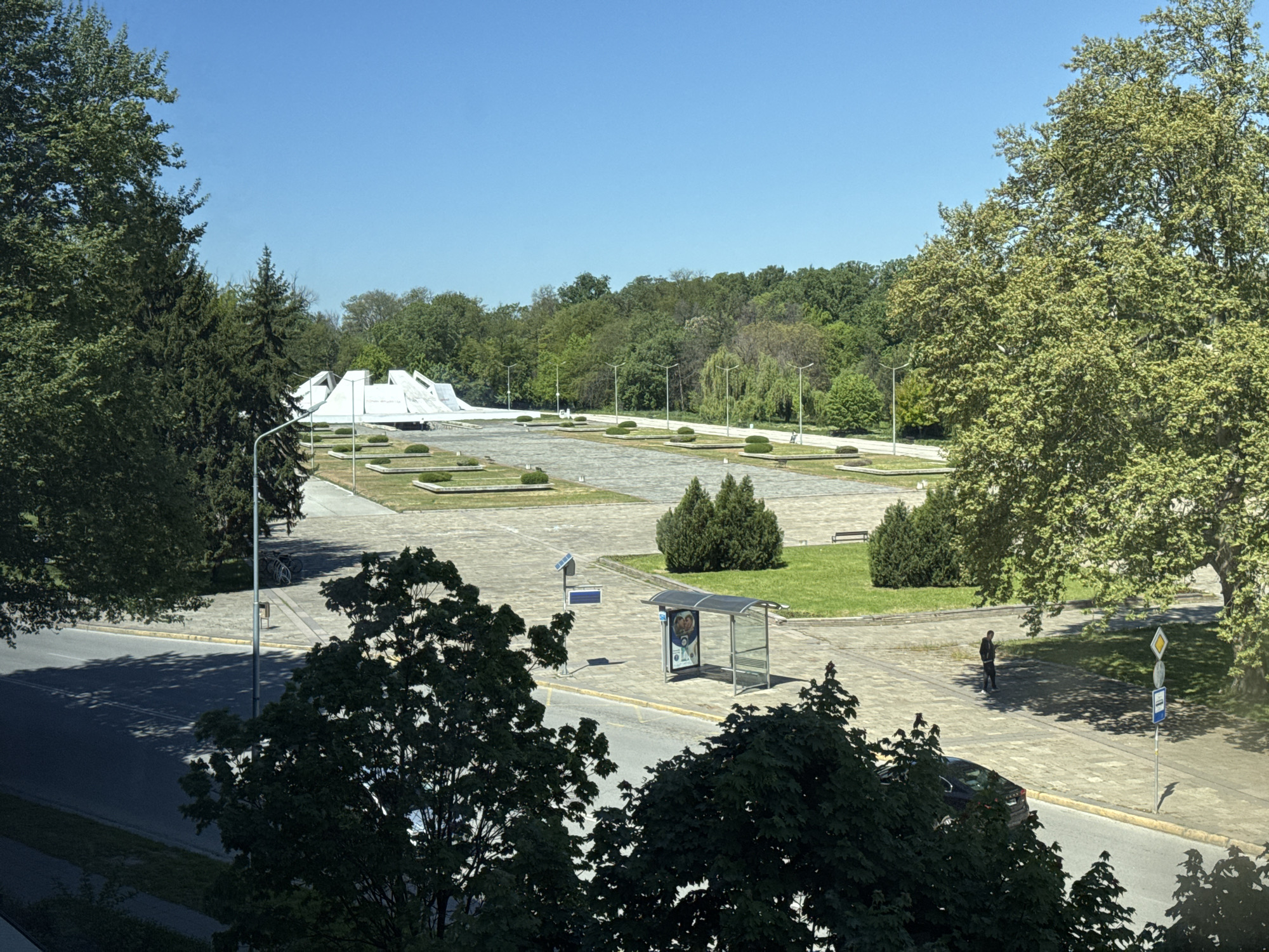 A clear daytime shot shows an expansive concrete plaza set against a backdrop of lush trees under a blue sky. In the distance, a white architectural structure with geometric shapes sits prominently in the center of the plaza. Manicured greenery dots the plaza surface. Closer to the viewer, a bus stop shelter stands along a street, with trees obscuring a full view of the road. A person is walking in the plaza. A car is parked near the street, partially hidden by trees in the foreground. Street signs are visible. The location appears to be an urban park or public space. The city and exact location are Unknown.