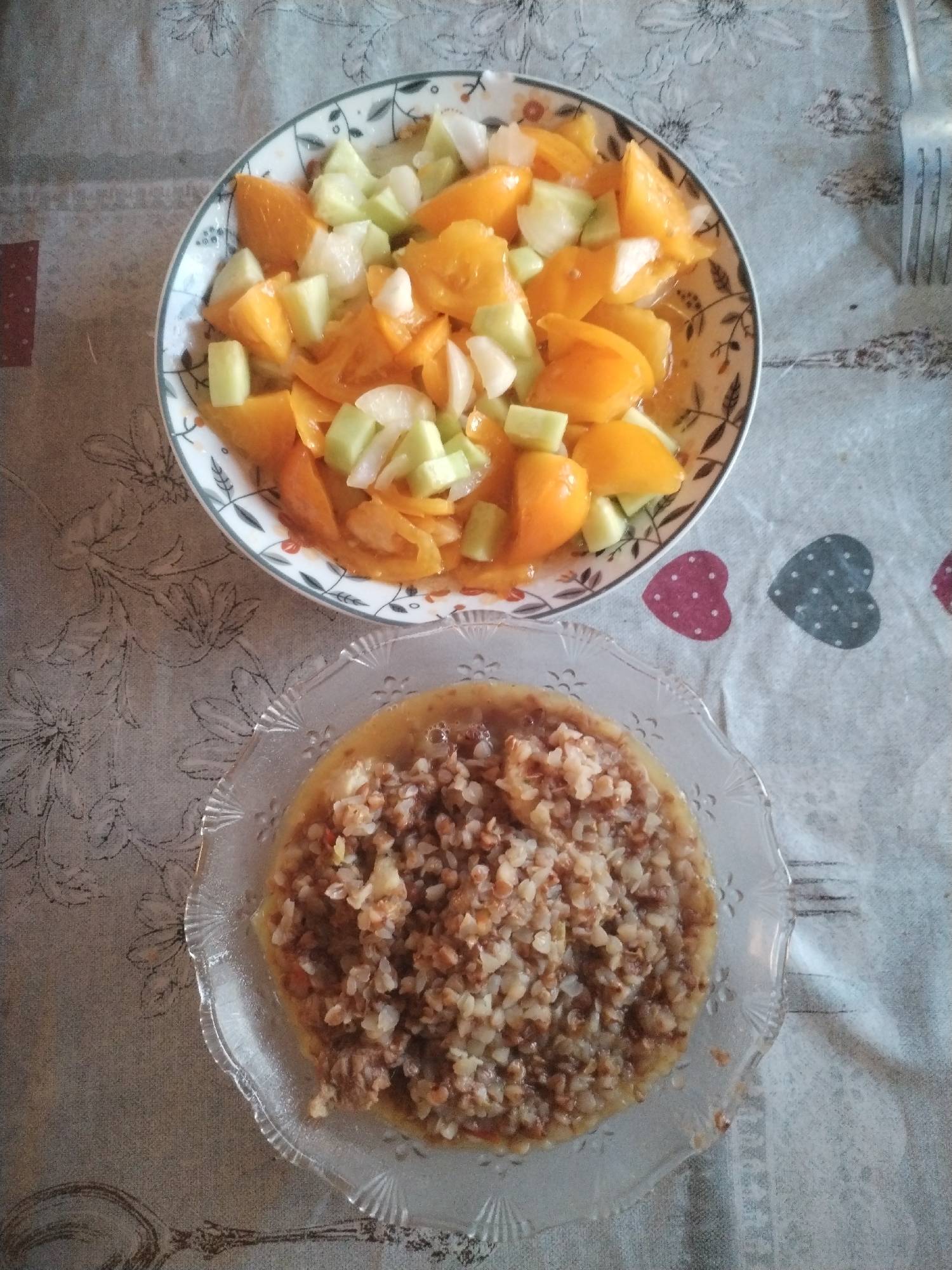 Two plates of food are displayed on a tablecloth. The top plate contains a salad of chopped yellow tomatoes and cucumbers. The bottom plate holds a portion of cooked buckwheat, possibly mixed with vegetables or meat. The tablecloth has a floral pattern and some decorative hearts. A fork is partially visible on the right side of the image. The setting appears to be indoors, likely a dining area.