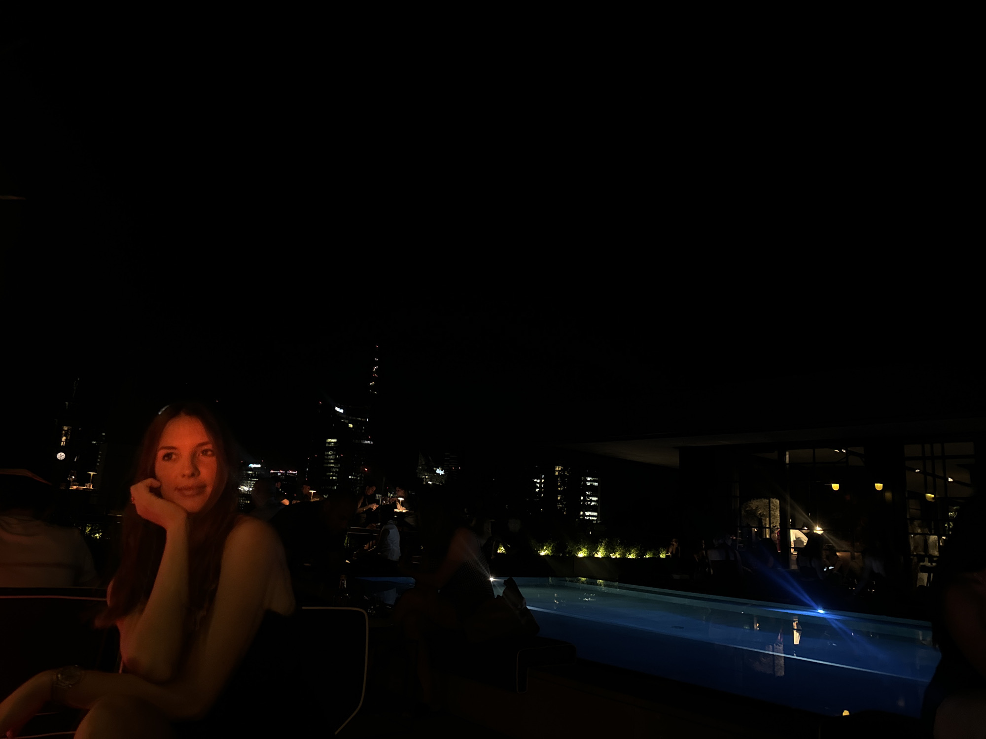 This is a nighttime, outdoor, long-shot photograph taken from a rooftop venue in Milan, Italy. A young woman with long brown hair, illuminated by warm light, is seated at a table on the left side of the frame, resting her chin on her hand and looking to her right. She appears to be wearing a dark, strapless dress and a watch on her left wrist. In the background, a swimming pool reflects blue light, and the illuminated skyline of Milan can be seen with numerous tall buildings and lights. Other patrons are visible in the background, some seated at tables and others blurred by motion, suggesting a social gathering or restaurant setting. The overall ambiance is dark with strategically placed lighting creating pockets of illumination.