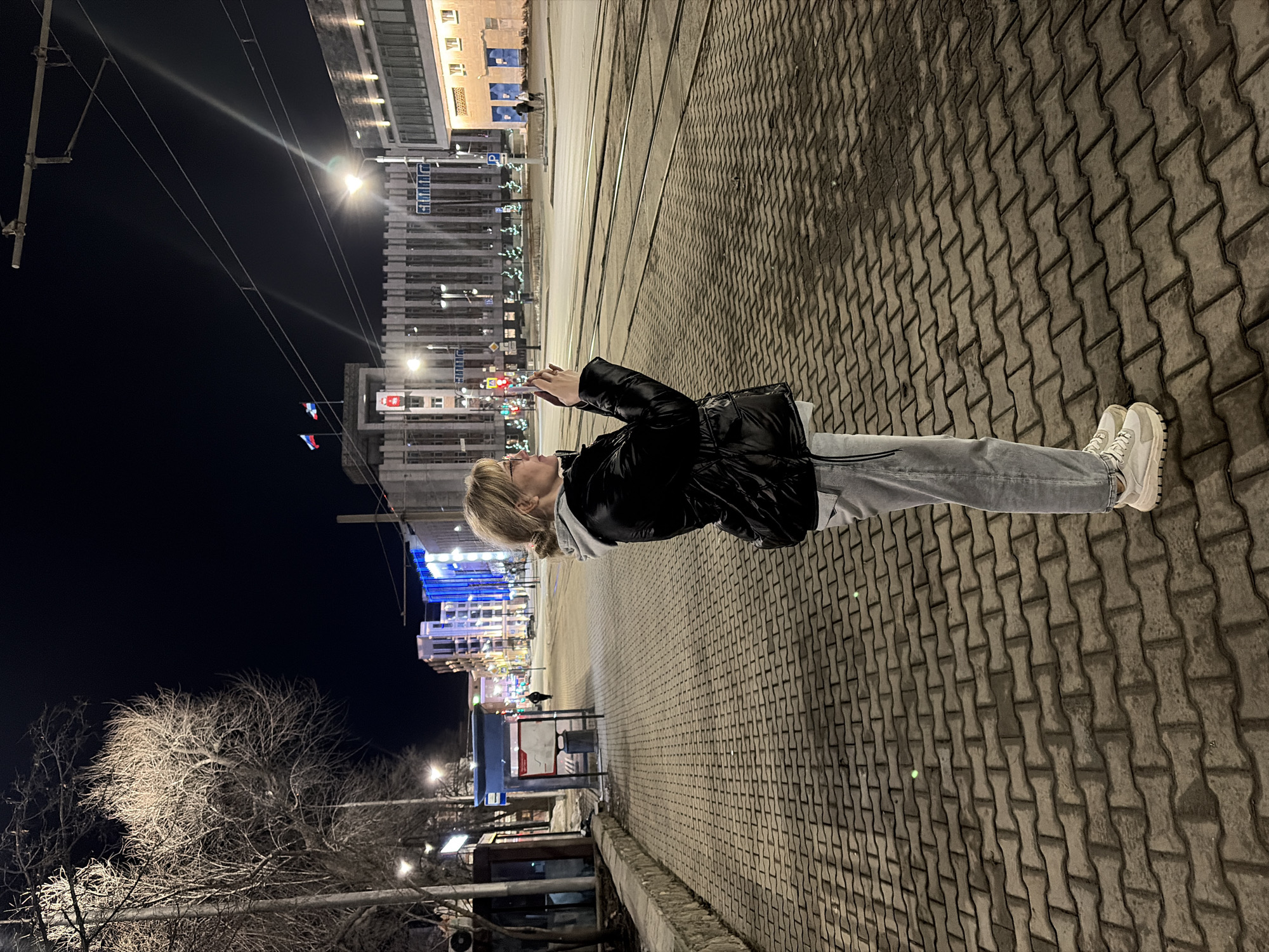 The image depicts a nighttime urban street scene on Lenina Street in Perm, Russia. A blonde female, with hair tied back and wearing glasses, stands in profile on a paved sidewalk. She is dressed in a black shiny puffer jacket over a light grey hooded sweatshirt, light grey jeans, and white chunky sneakers. She holds a smartphone horizontally with both hands, aiming it towards the right, appearing to photograph the illuminated cityscape.
To the left of the subject, a low stone curb separates the sidewalk from bare-branched trees and a utility pole. Further left, a blue-roofed bus stop shelter with a timetable and a trash bin is visible. The sidewalk, constructed from grey interlocking pavers, extends into the foreground. Adjacent to the sidewalk runs a street containing two sets of tram tracks, with the surface appearing slightly soiled or lightly covered.
In the background, directly across the street, stands the multi-story Legislative Assembly of Perm Krai building, located at Lenina Street, 51. This grey building features prominent vertical architectural elements. Two flags fly from its rooftop: the Russian Federation flag and the Perm Krai flag. A red emblem, identified as the Perm Krai coat of arms (depicting a bear with a book), is centrally displayed on the building's facade. Streetlights, overhead tram/trolleybus wires, and illuminated building facades, some with blue accent lighting, define the scene against a dark night sky. Several distant pedestrians are visible further down the street. The bare trees suggest a late autumn or winter season.