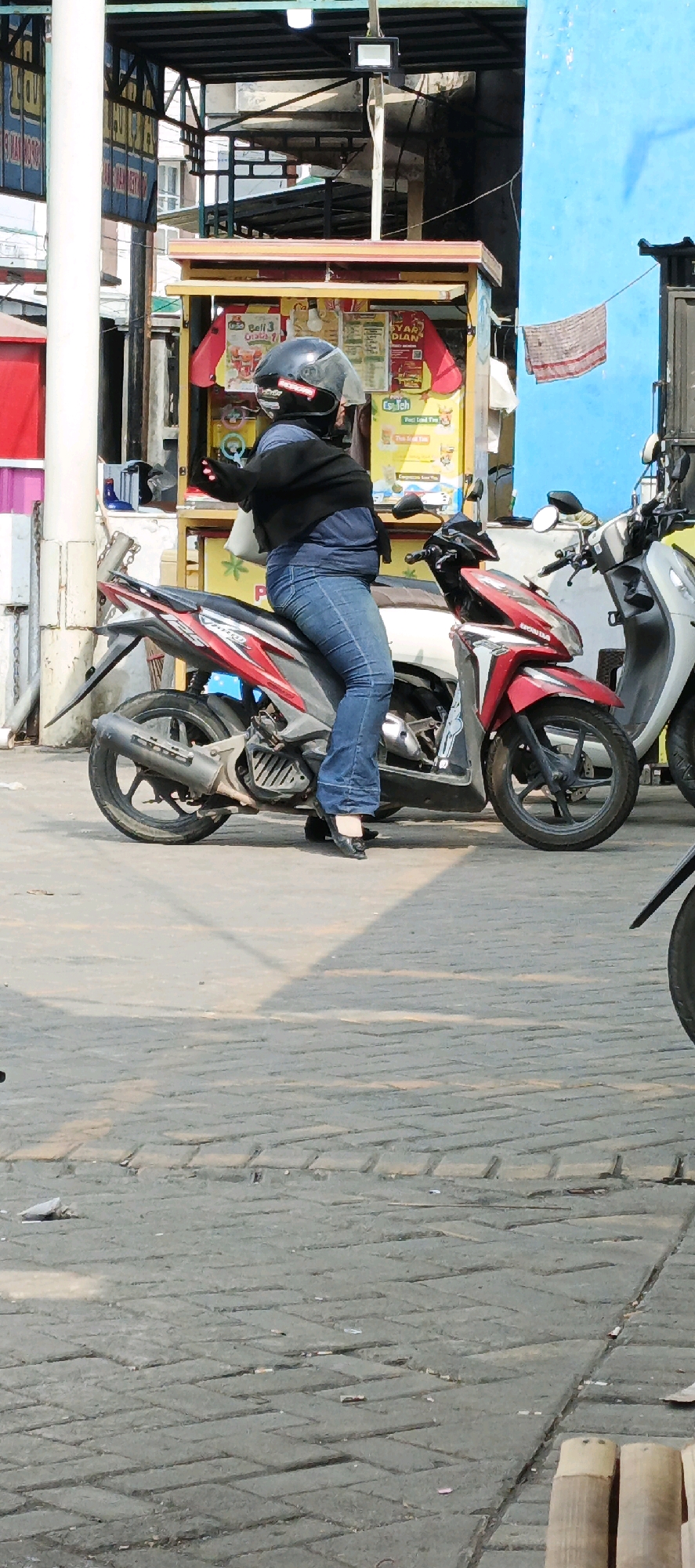 A person wearing a helmet and black jacket over a blue shirt and blue jeans is seated on a red and gray scooter in Kasiyan, Indonesia. The person's left hand is visible, extended with gloved fingers. The scooter is parked on a paved area with interlocking gray bricks. Behind the scooter, a blue wall is visible, with a red and yellow kiosk in the foreground. The kiosk has various signs with Indonesian text and images of drinks. To the right of the main scooter, a white scooter is parked. The environment appears to be an outdoor urban setting, possibly near a street or market.
