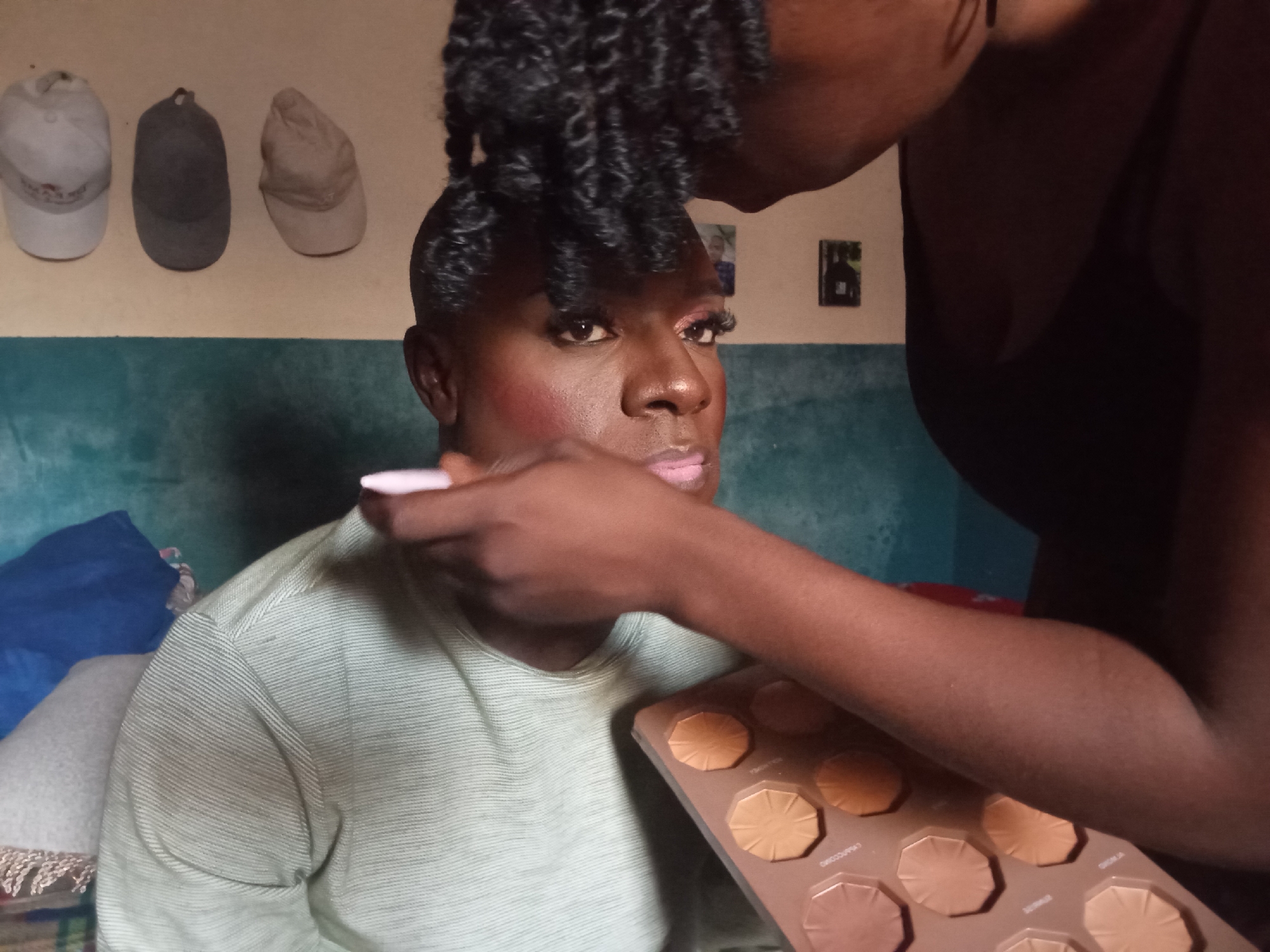 The image depicts a close-up of two individuals, likely in a residential setting, as indicated by the presence of wall decor and household items. The foreground focuses on a person applying makeup to another. The person receiving makeup is wearing a light green striped t-shirt. They have their eyes open and are looking towards the viewer. Pink blush is visible on their cheeks, and their lips are painted with a light pink lipstick. Long, false eyelashes are also applied. A makeup palette, containing various shades of foundation or contour, is held by the person applying the makeup, whose arm and hand are visible in the frame. The background shows a wall with three baseball caps hanging on it and two framed pictures. The person applying makeup has dark, curly hair styled upwards. The scene appears to be indoors, with a teal-colored wall or surface dominating the background. The overall lighting suggests an indoor environment, possibly a home. The location is identified as Rukuba Road, Nigeria.