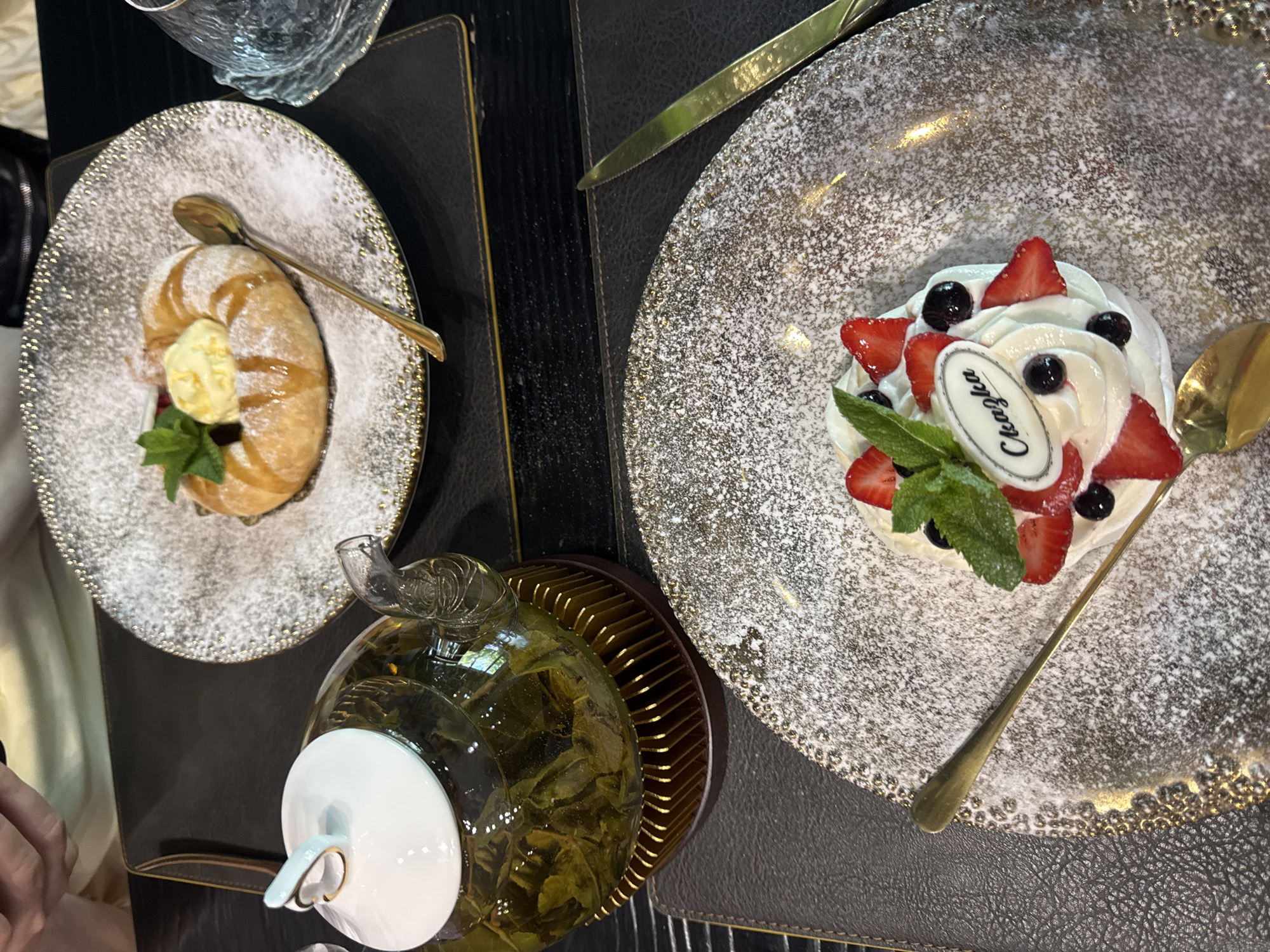 In this overhead shot, two elegantly plated desserts sit on dark placemats, next to a glass teapot filled with green tea and a person's hands in the upper left corner. The dessert in the foreground is a white meringue topped with whipped cream, sliced strawberries, blueberries, and mint leaves. A small oval label with black text that reads "Сказка" is affixed to the cream. A golden spoon rests on the plate.
The dessert in the background is a circular pastry with a hole in the center, filled with a scoop of pale yellow ice cream and topped with mint. Drizzled with a golden syrup, it is also dusted with powdered sugar. A golden spoon is placed on this plate as well. A tea glass sits to the right of this dessert. The scene suggests a restaurant or cafe setting.