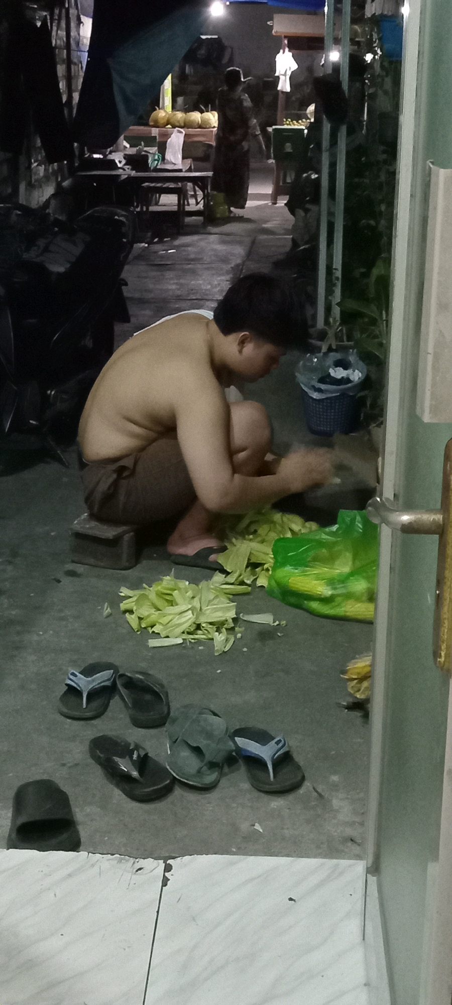 A person is crouched on the ground in what appears to be a market or an outdoor area, processing vegetables, likely corn husks. The person is shirtless and wearing brown shorts. A pile of cut vegetable matter, which appears to be corn husks, is spread out on the ground in front of them. To their right is a green plastic bag also containing corn. Multiple pairs of flip-flops are scattered around the ground. In the background, a woman in a long dress is visible near a stall displaying several yellow gourds. The scene is dimly lit, suggesting it is either evening or an indoor/covered area. The overall environment seems to be part of a marketplace in Babat, Indonesia, given the presence of fresh produce and the general atmosphere.