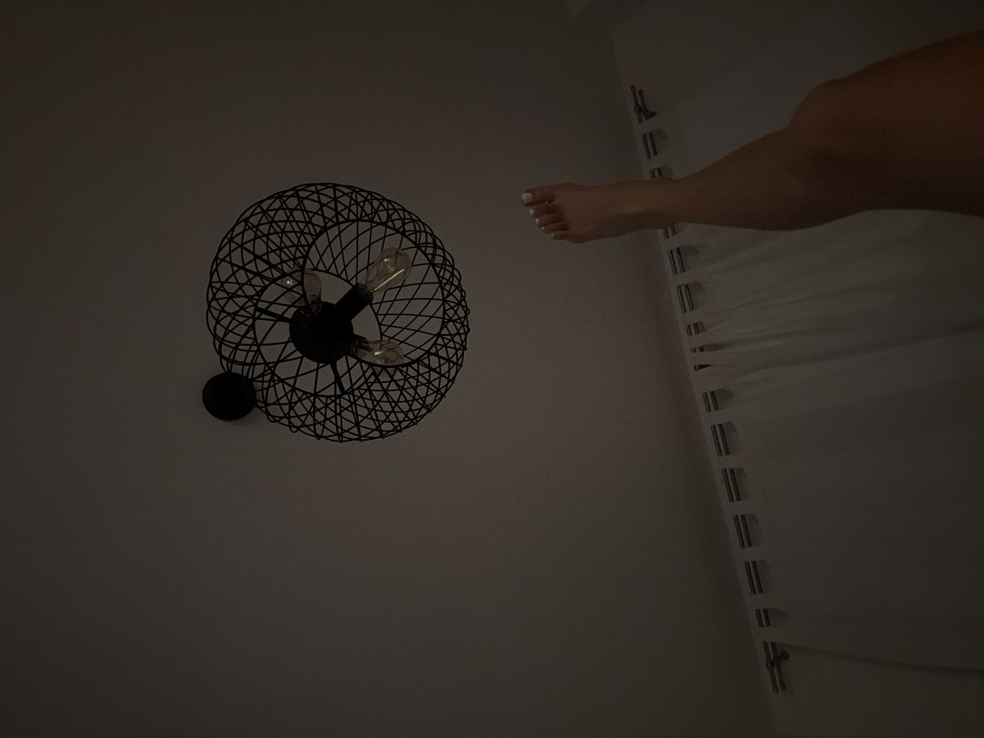 A low-angle shot shows a dark room in Plovdiv, Bulgaria. A black, round, wire-frame light fixture with exposed light bulbs is on the left wall. To the right, a portion of a person's leg and foot extends from behind what appears to be a white curtain or sheet hanging from curtain hooks. The person's toenails are painted white. The overall lighting is dim.