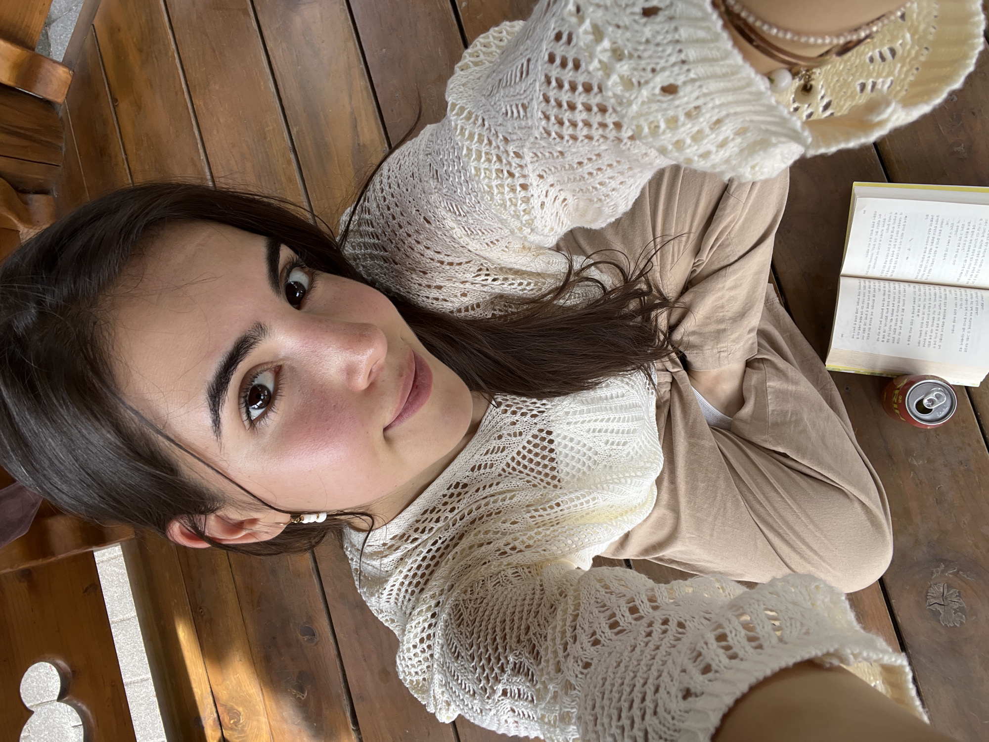 A young woman with dark hair is sitting cross-legged on a wooden deck, looking up at the camera. She is wearing a cream-colored, crocheted, bell-sleeved sweater and tan pants. She has freckles on her nose and cheeks and is wearing a delicate earring in her left ear. Next to her, on the wooden floor, is an open book and a red soda can. The wooden deck appears to be part of an outdoor seating area, possibly at a cafe or park in Daegu, South Korea.