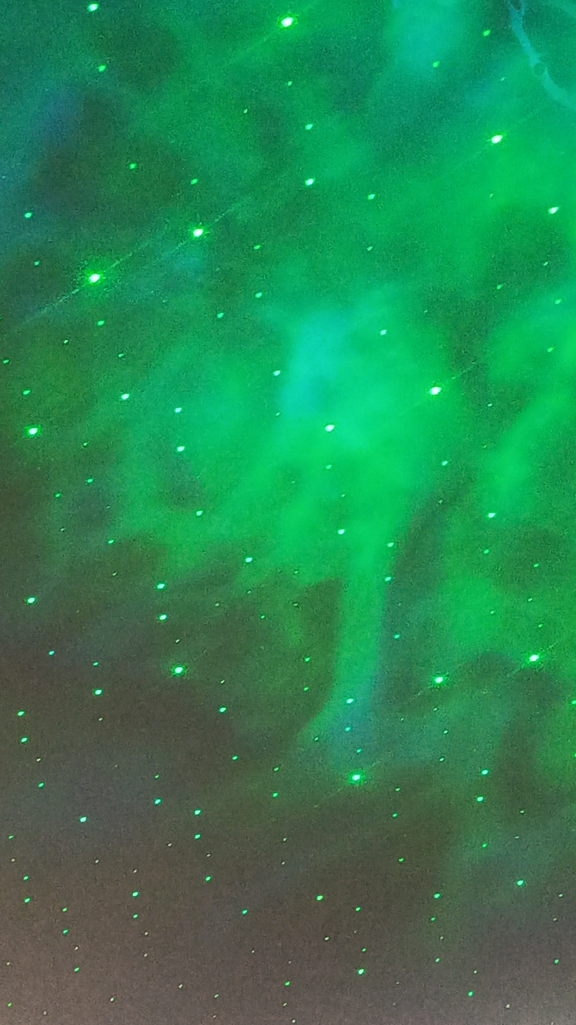 The image depicts a dark background illuminated by scattered green lights, resembling a starry sky or a galaxy projection. There are no people or identifiable objects present. The lighting creates a hazy, ethereal effect with areas of brighter green glow and dimmer, more diffused light. It's impossible to determine the specific location in Zvenigorod, Russia, from this abstract imagery.
