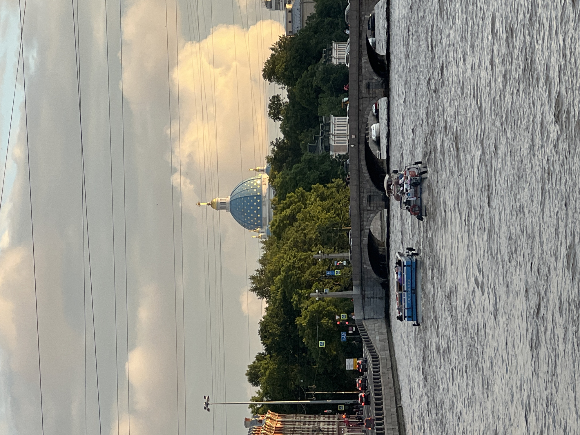 This image depicts a scene in Saint Petersburg, Russia, likely along the Neva River. In the background, a prominent blue dome with golden accents, possibly part of a church or administrative building, is visible above a line of lush green trees. A stone bridge with multiple arches crosses the river, and traffic, including cars, is moving on the embankment to the left. The foreground shows the choppy surface of the river, with two boats carrying passengers navigating the water. The sky is partly cloudy with soft light, suggesting it is either morning or late afternoon. The overall atmosphere is calm and urban.