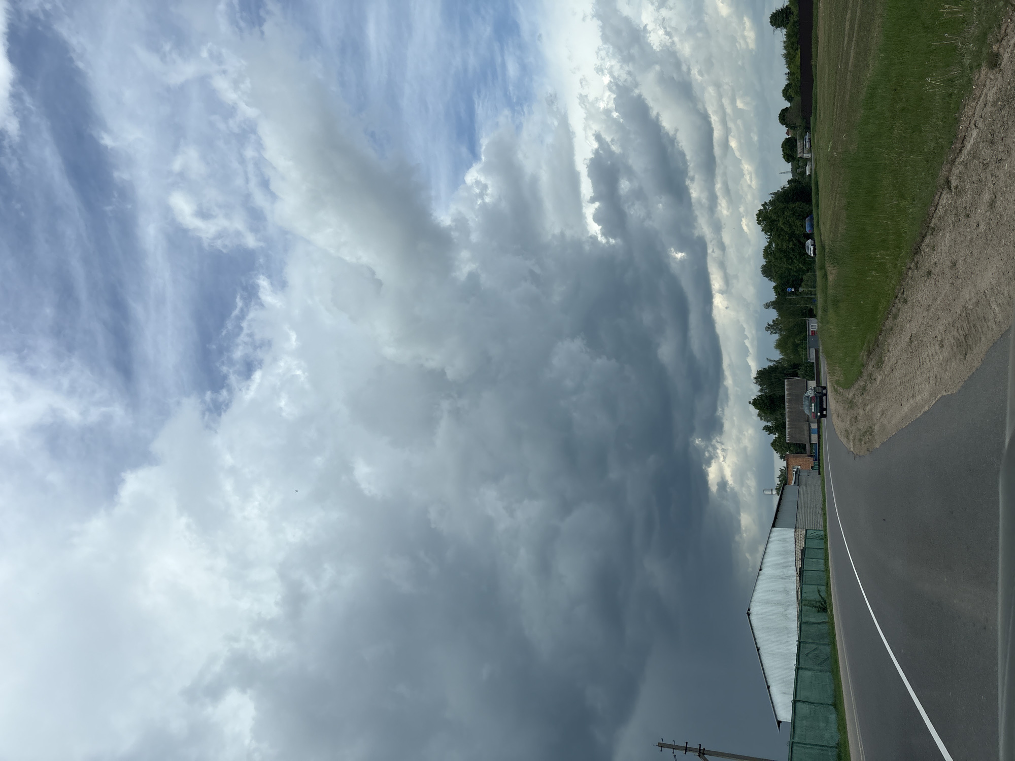 A photograph taken in Голацкі сельскі Савет, Belarus, depicts a roadside scene under a sky dominated by dramatic, dark storm clouds. A two-lane asphalt road curves gently, marked by a solid white line separating the lanes. Adjacent to the road is a grassy verge transitioning into a field. On the left side of the road, structures, including a long, low building with a light-colored corrugated roof and green fencing, are visible. Power lines are present. The sky features a mix of blue patches and dense, gray clouds.