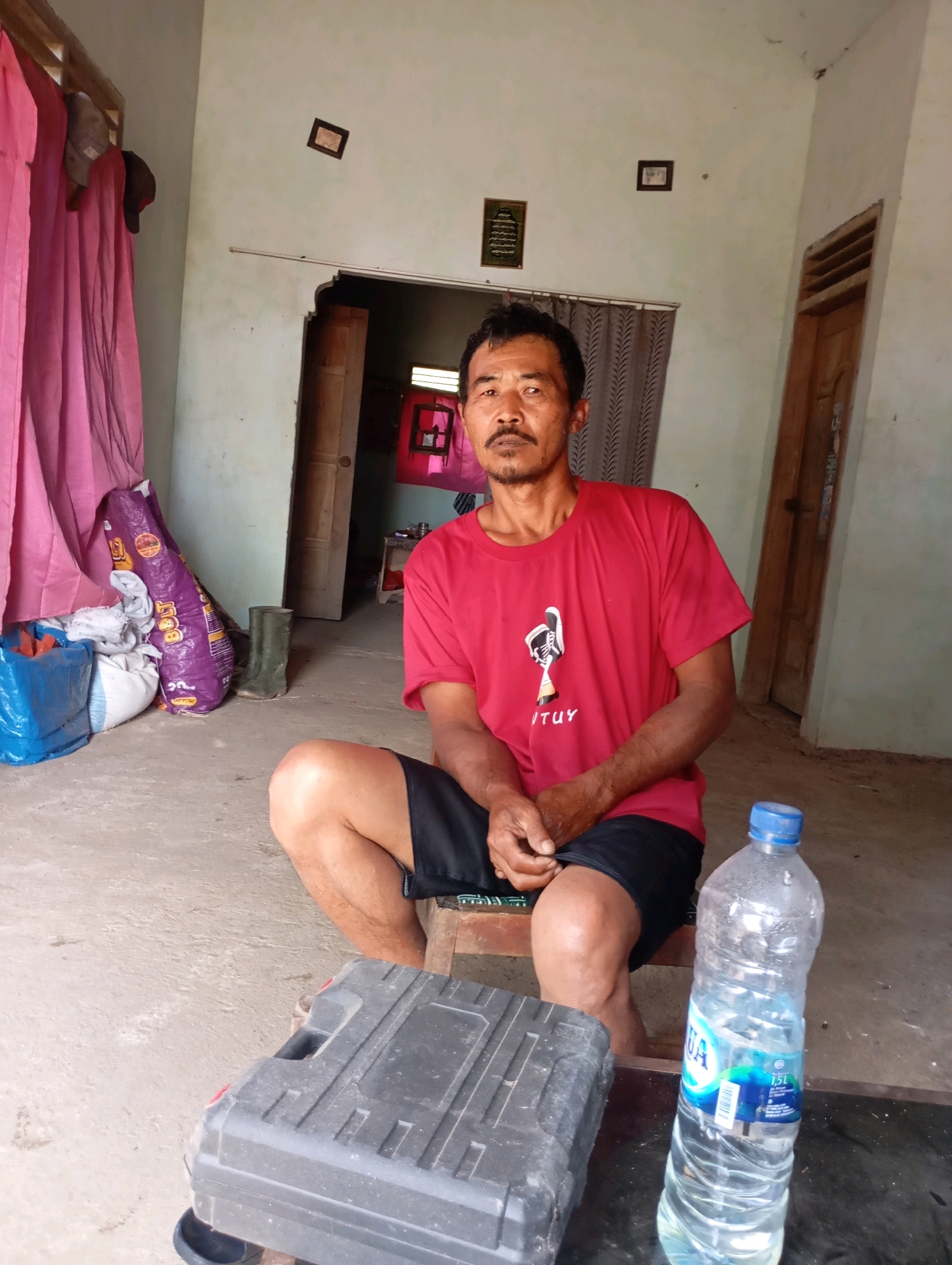 In the city of Ketepung, Indonesia, a man with dark hair and a mustache sits on a stool in what appears to be a home. He is wearing a red t-shirt with a graphic print and black shorts. In front of him, on the floor, are several items including a black toolbox, a clear plastic bottle of water, and to his left, a purple bag and other bundles of goods. The room has light-colored walls and a doorway in the background. There are also two framed pictures on the wall.