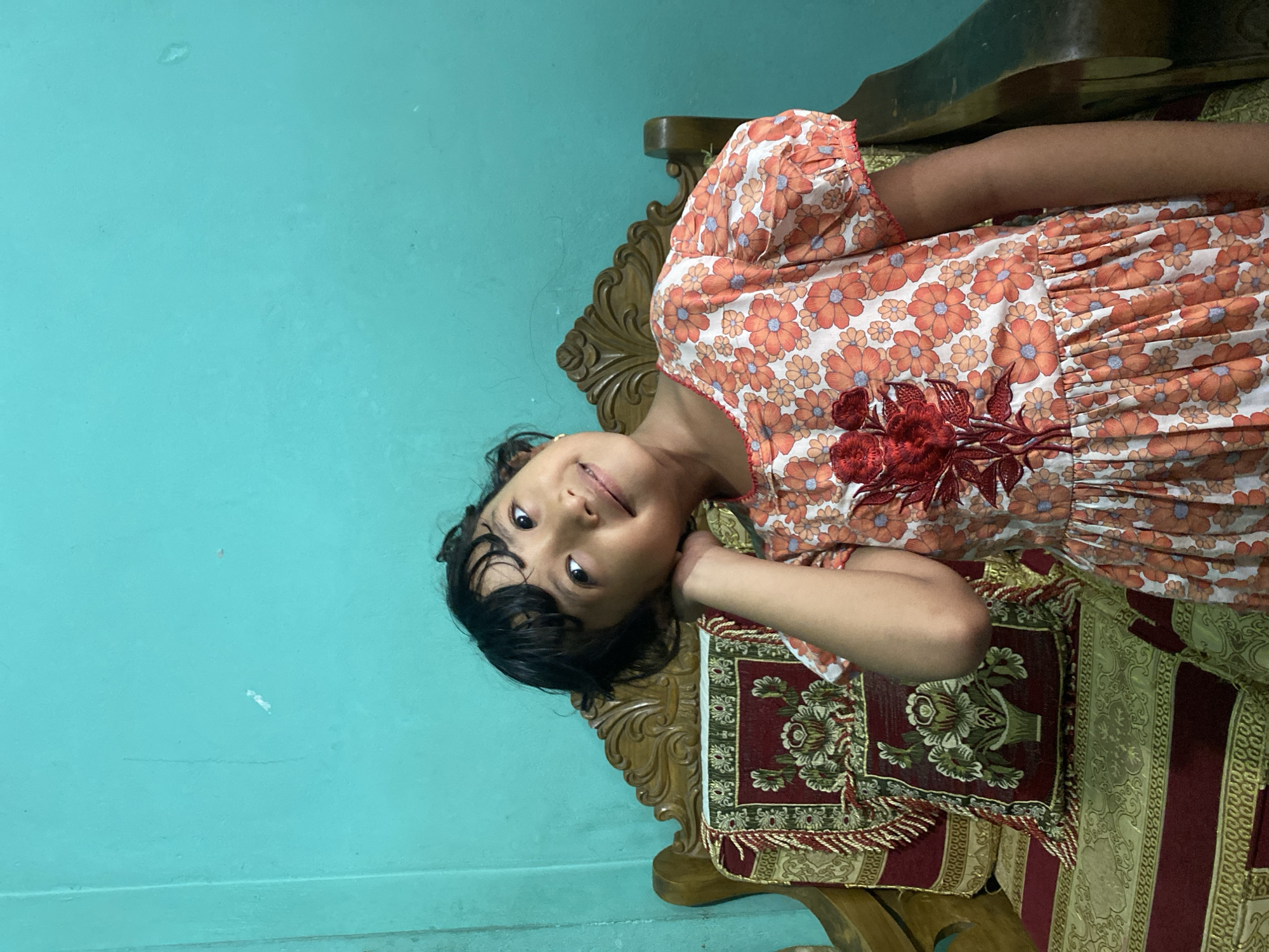 A young girl lies on a carved wooden chair, possibly in Dhaka, Bangladesh. She wears an orange and white floral dress with a red embroidered design. The chair is covered in a patterned fabric with red and gold tones. The background is a turquoise wall. The girl's head rests on the chair's arm, and she looks at the camera with a slight smile. Her hair is dark and slightly disheveled.