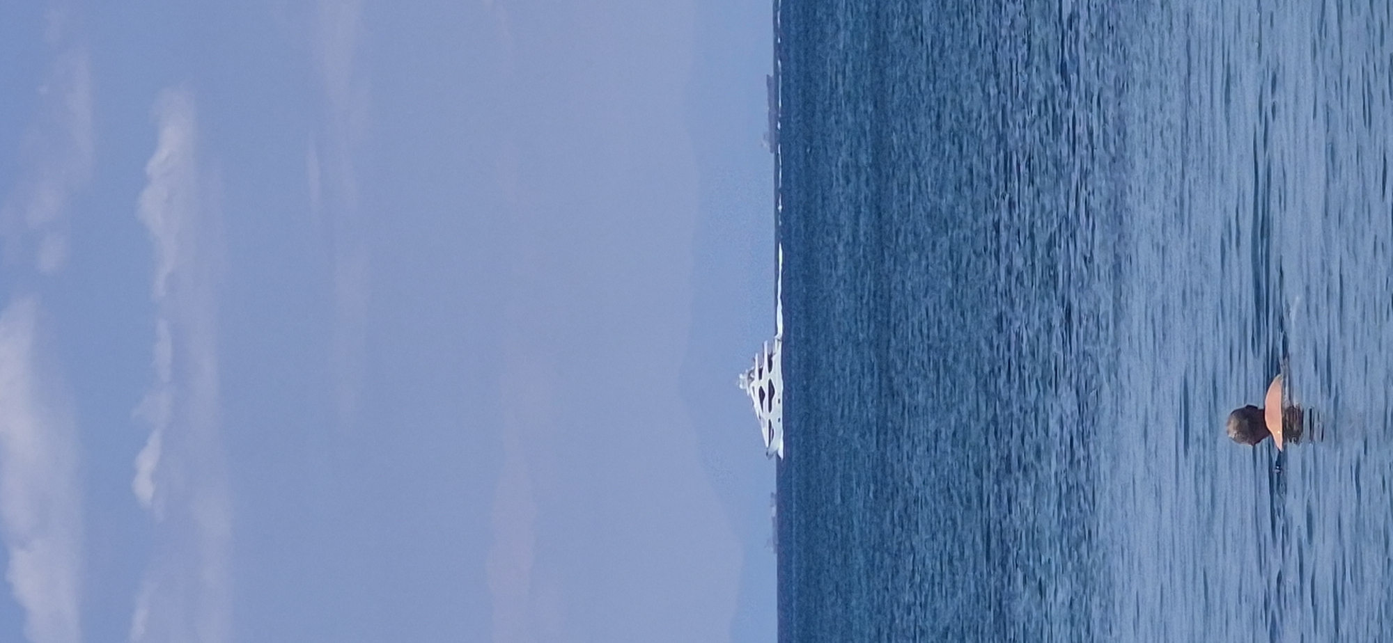 A bright, sunny day shot captures a wide expanse of blue sea, with gentle waves rippling on the surface. In the foreground, a person with blonde hair is partially submerged in the water, facing away from the camera. Further out, a large, white luxury yacht navigates the sea, leaving a white wake behind it. In the distance, hazy mountains form a backdrop against a pale blue sky with scattered clouds. The scene is consistent with a view from the coast of Paleo Faliro, Greece, looking out towards the Saronic Gulf.